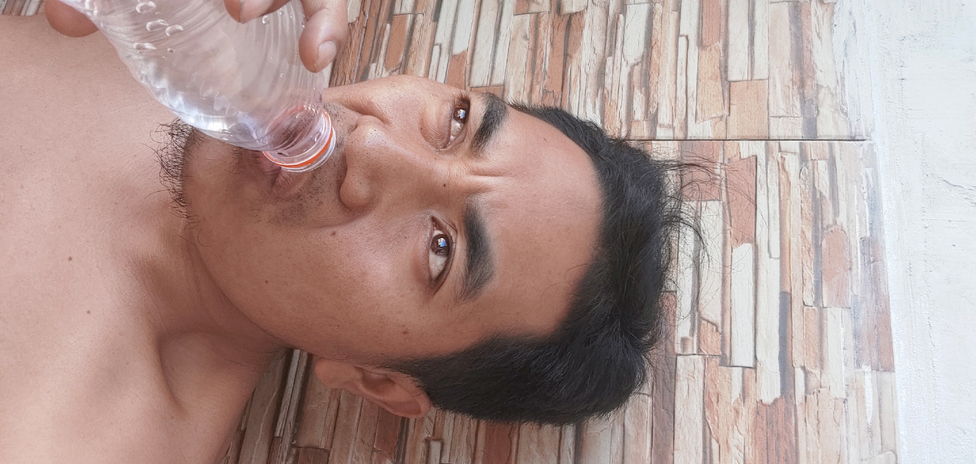 In Kembang Kerang, Indonesia, a close-up shot captures a man drinking from a clear plastic water bottle. The man, with dark, tousled hair and visible stubble, is positioned against a textured wall composed of rectangular stone-like tiles in shades of brown, beige, and white. His eyes are looking upwards and to the left, his brow furrowed as he drinks. The man's bare shoulder and upper chest are visible. The scene is lit with natural light, highlighting the details of his skin and the texture of the wall.