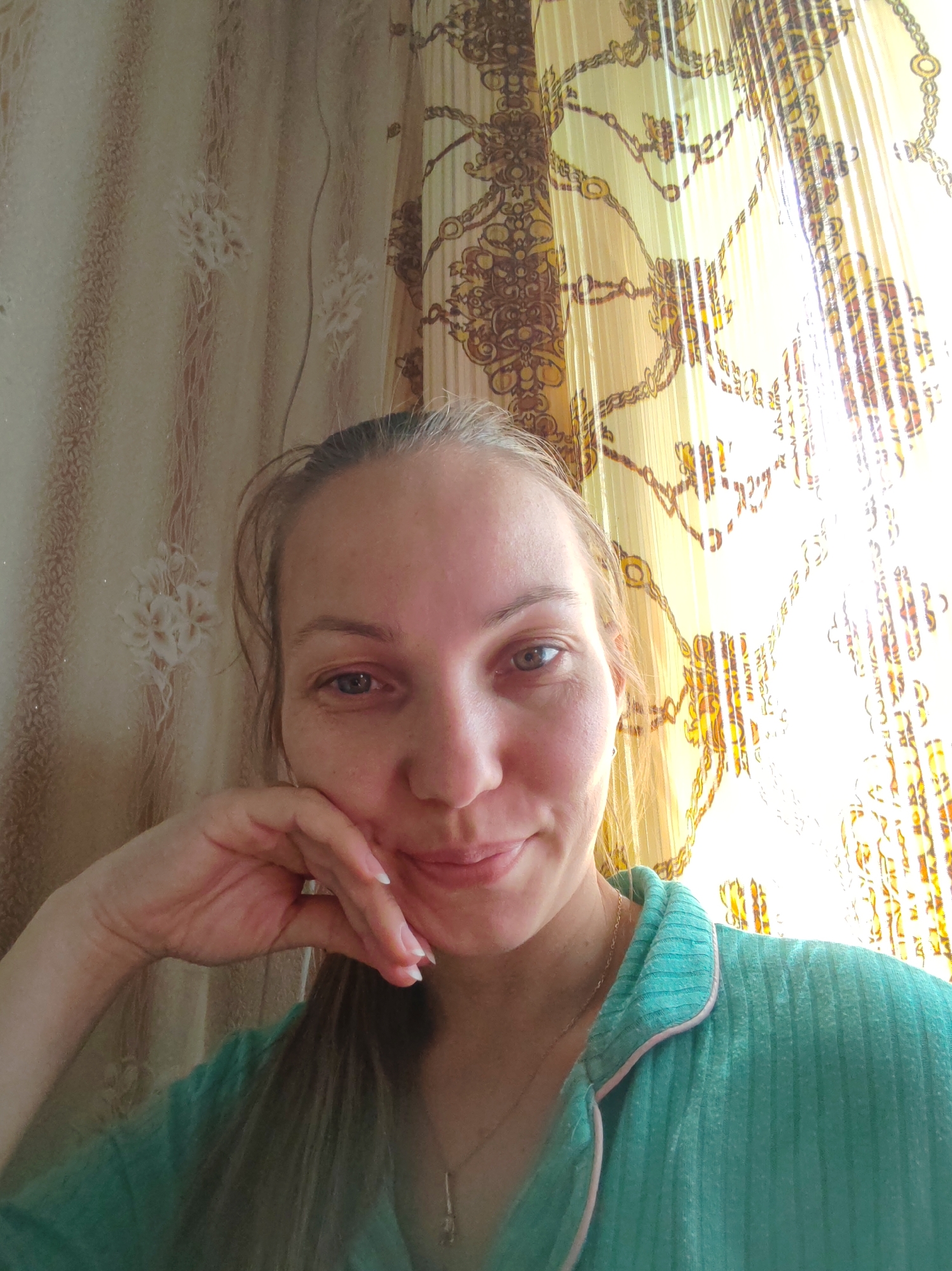 A woman is taking a selfie indoors, possibly in a bedroom. She is leaning her head on her hand and smiling faintly. She is wearing a teal pajama top with pink trim. The background consists of two types of curtains. The left curtain is a light-colored fabric with a subtle floral pattern, while the right curtain is a sheer, golden material with a large, ornate chain-link pattern. Sunlight is streaming through the right curtain, creating a bright and somewhat washed-out effect in that area. The image does not contain any information about the city of Чухлэм, Russia.