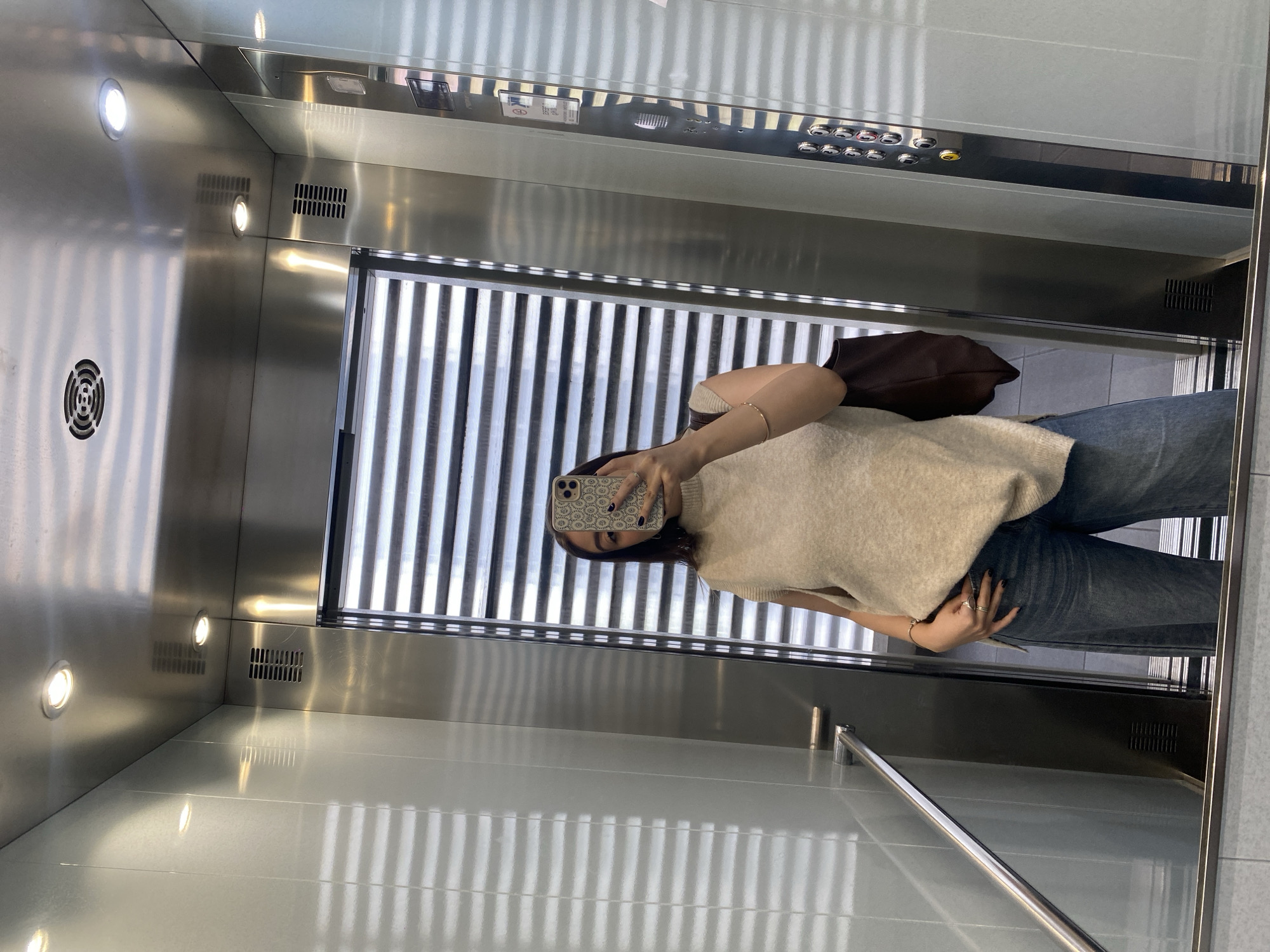 A woman is taking a selfie in an elevator. The elevator has a metal interior with vertical striped panels on the back wall. There are lights recessed into the ceiling and a control panel above the door. The woman is wearing a cream-colored sweater vest and blue jeans. She is holding her phone in front of her face to take the picture. A brown purse is also visible in the background. The location appears to be an interior, possibly an office building, but the city is Unknown.
