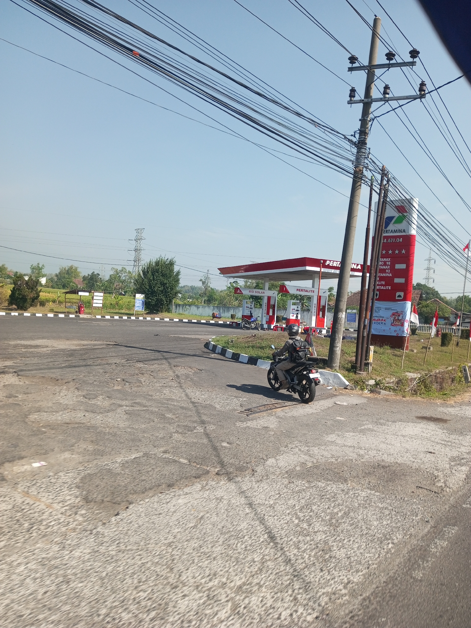 The image shows a Pertamina gas station in Pucangsari, Indonesia. A person is riding a motorcycle on a paved road in front of the station. The gas station has red and white signage and multiple pumps. There are power lines running overhead and a utility pole with ceramic insulators. Trees and vegetation are visible in the background, along with a tall transmission tower in the distance. The sky is clear and blue.