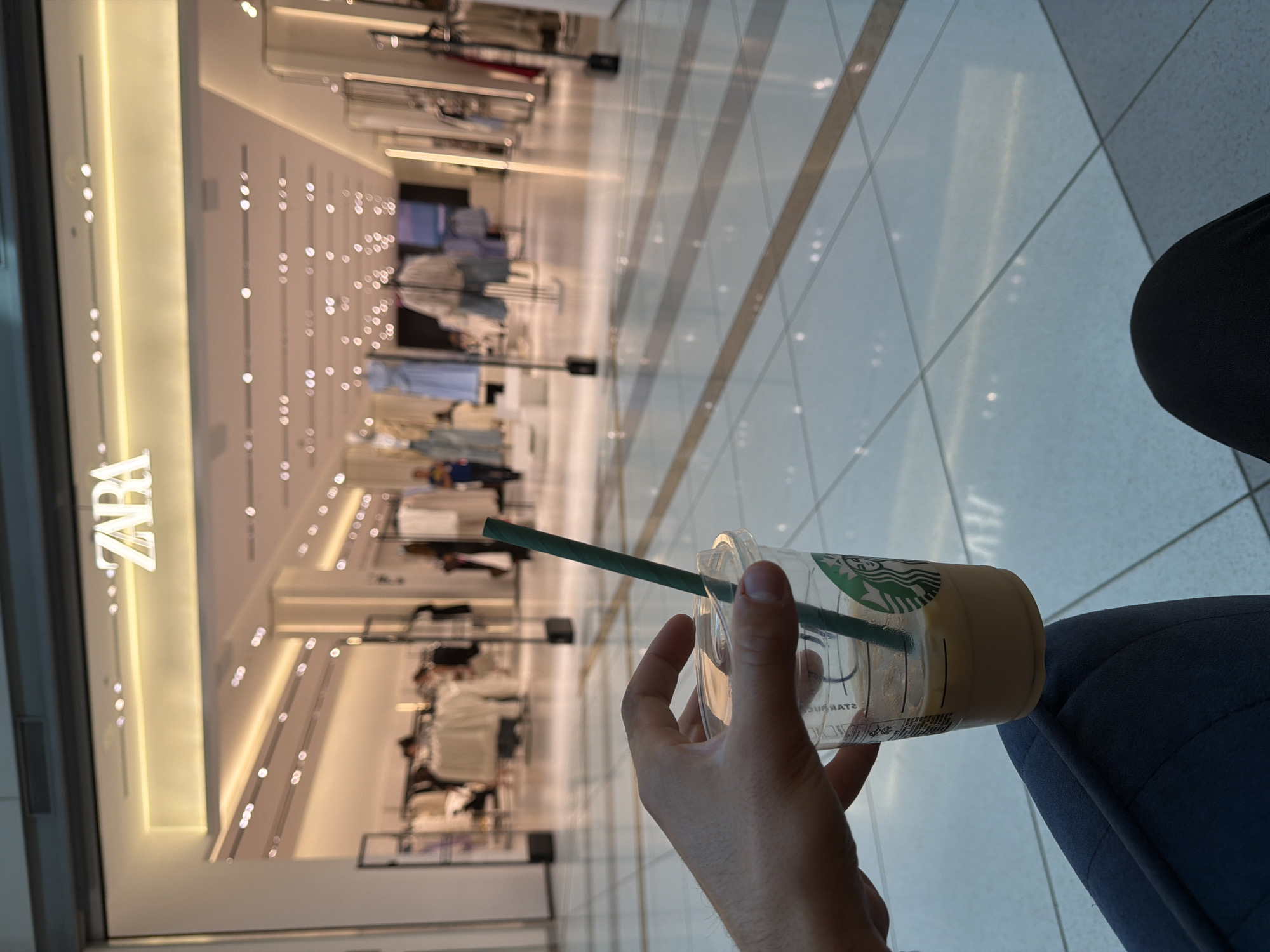 The image shows a person's hand holding a Starbucks cup with a green straw, near their lap which is covered by dark blue fabric, possibly a chair or clothing. The cup is partially filled with a light-colored beverage. In the background, a brightly lit store interior is visible, with clothing racks and displays. The store appears to be Zara, as evidenced by the visible text "ZARA" on a sign. The flooring in the background is tiled and reflective, suggesting a modern indoor environment like a shopping mall. The scene is captured from a slightly upward angle. The lighting suggests it could be daytime, possibly indoors with artificial lighting. There are no clear indications of weather conditions. The image seems to capture a moment of personal time, perhaps a break, in a commercial setting.