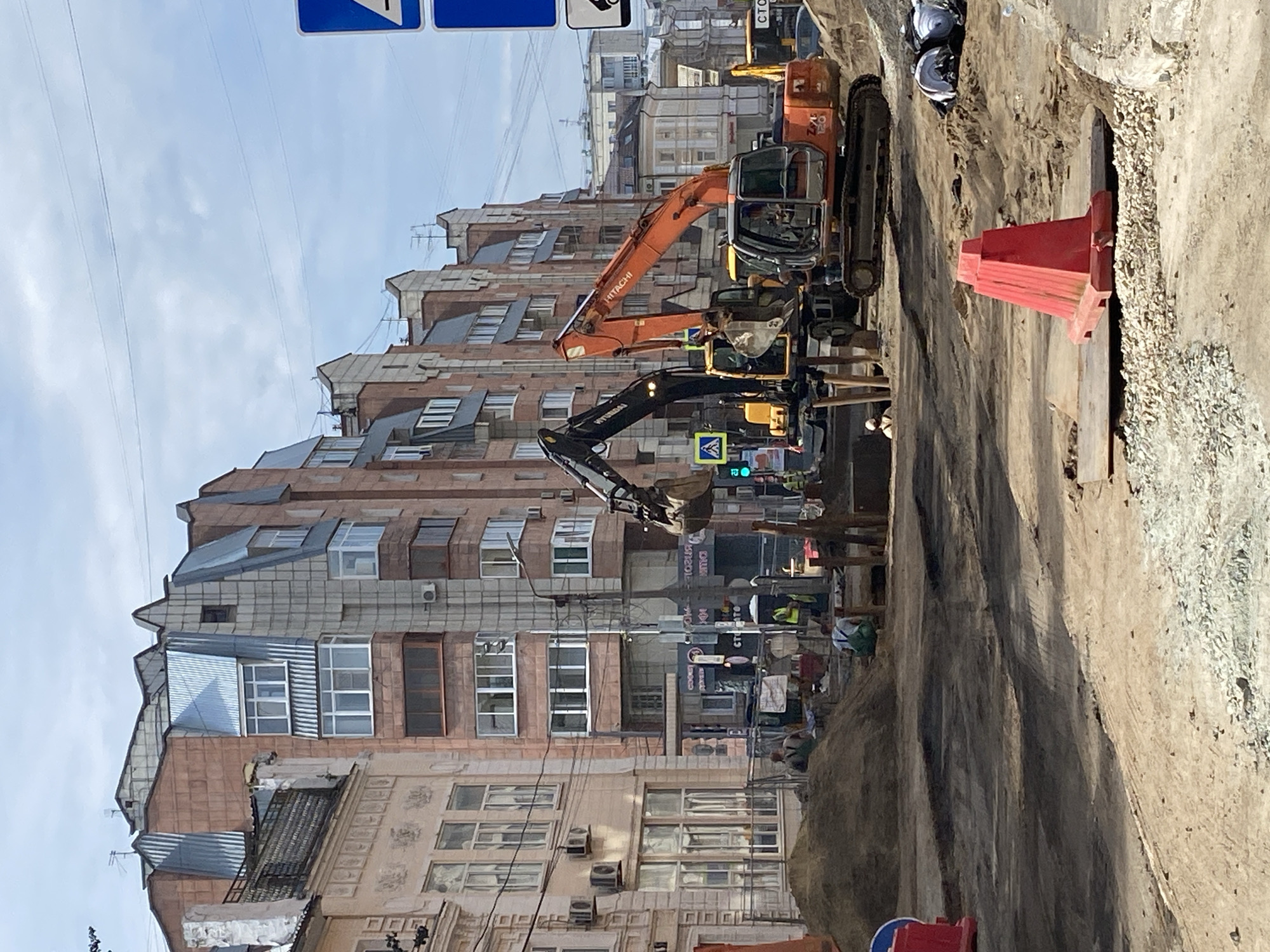 The image depicts a construction site on a city street in Perm, Russia. In the foreground, disturbed earth and gravel indicate ongoing work, with a prominent red traffic barrier and a metal plate. Several construction vehicles, including two excavators (one orange Hitachi and one black Hyundai), are positioned on the site. Workers in high-visibility vests are present, engaged in various tasks. The background features a multi-story building with a distinctive mansard roof and several windows. Overhead, power lines are visible against a cloudy sky. The scene suggests a street reconstruction or utility work in progress within an urban environment.