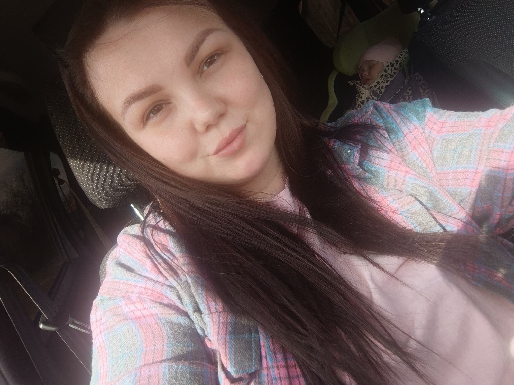 A selfie photograph taken in what appears to be the interior of a car. A woman with long, dark hair is the primary subject; she wears a pink t-shirt under a blue and pink plaid shirt. In the background, there is a child asleep in a car seat wearing a pink hat. The background suggests the interior of a vehicle with dark upholstery. It is not possible to confirm the location in Chaikovsky, Russia.