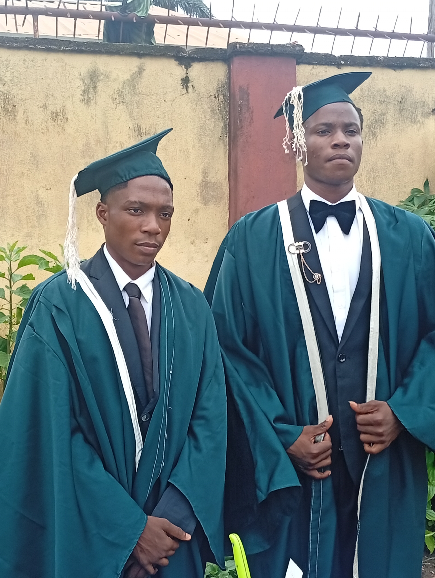 Two young men, likely students, stand outdoors wearing graduation regalia. Both are dressed in dark suits with white shirts and ties (one a black tie, the other a black bow tie). They are covered in dark teal graduation gowns, which are draped over their shoulders and arms. Each man also wears a dark teal graduation cap with a white tassel. The man on the left has his hands clasped in front of him, while the man on the right has his right hand resting on the lapel of his gown and his left hand is partially obscured. They are standing in front of a textured, pale yellow wall, with some greenery visible to the left and right. A weathered metal fence with pointed uprights is visible at the top of the frame, against a pale sky. The lighting suggests it is daytime.