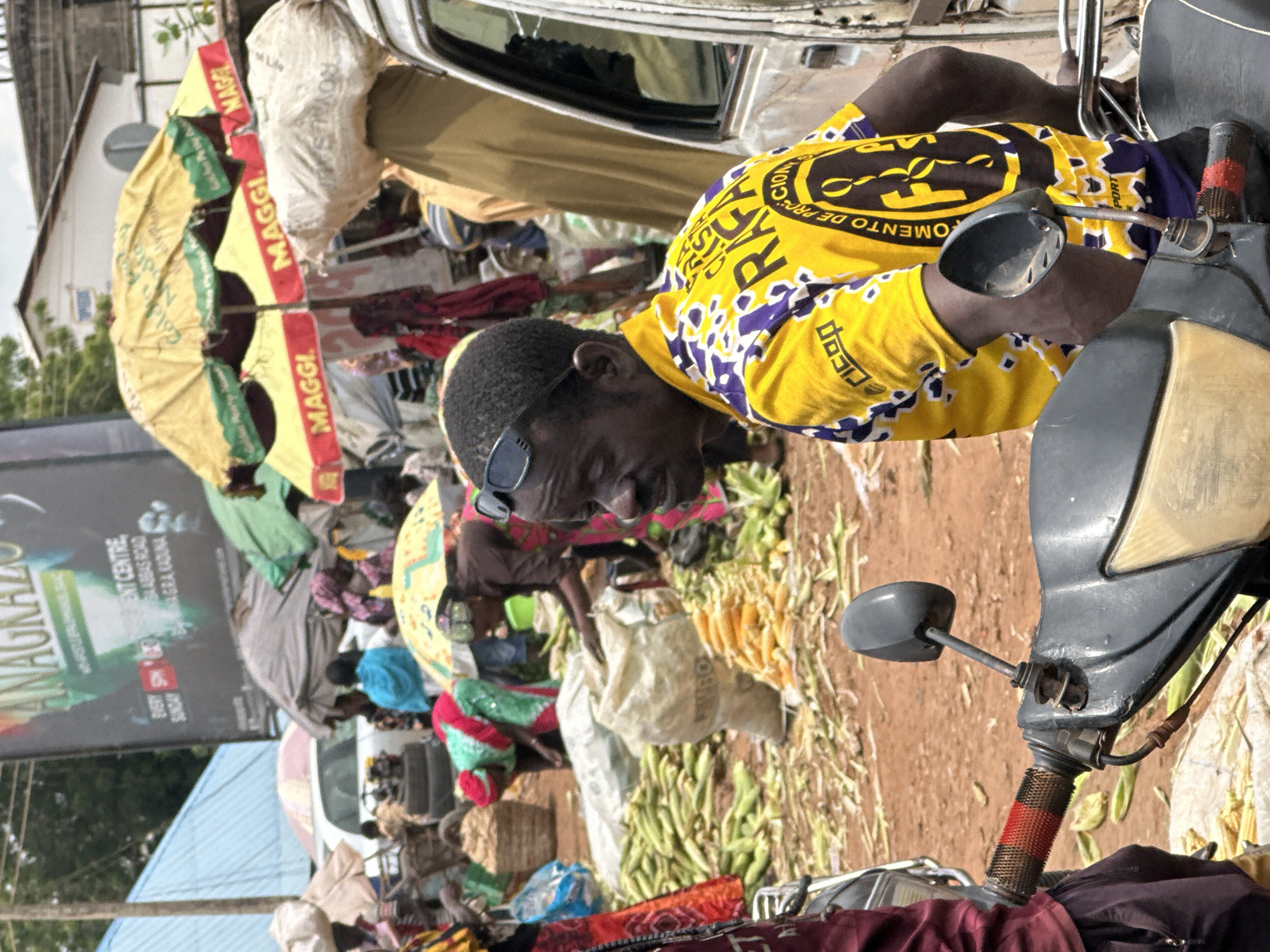 The image depicts an outdoor market scene in Sabon Tasha, Nigeria. In the foreground, a man wearing a yellow and purple patterned shirt is seated on a motorcycle. He has sunglasses pushed up on his forehead. Behind him, piles of corn are visible on the ground, along with large sacks. Several other individuals are present in the market, some tending to the goods or walking amongst the stalls. Various stalls are set up with umbrellas providing shade, some of which are branded with "MAGGI". In the background, there is a large billboard and buildings characteristic of a Nigerian urban setting. The ground is dusty and appears to be unpaved.