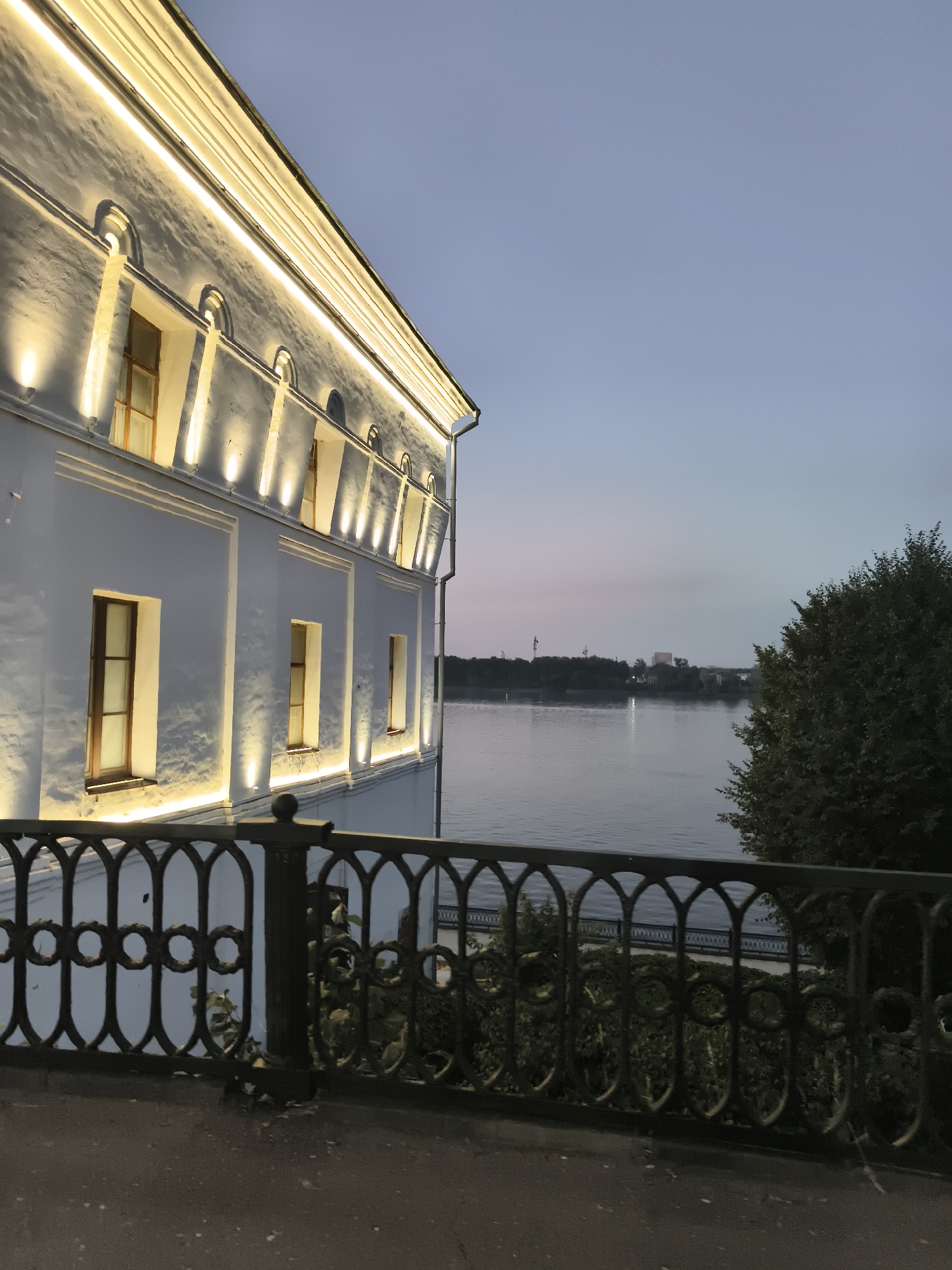 The image shows the illuminated facade of a white building with arched windows, lit by warm lights, against a twilight sky. In the foreground, a decorative black metal fence runs along a walkway. Beyond the fence, a wide river is visible, with trees and buildings on the opposite bank. The scene appears to be along the Volga River embankment in Yaroslavl, Russia, possibly near the Strelka confluence.