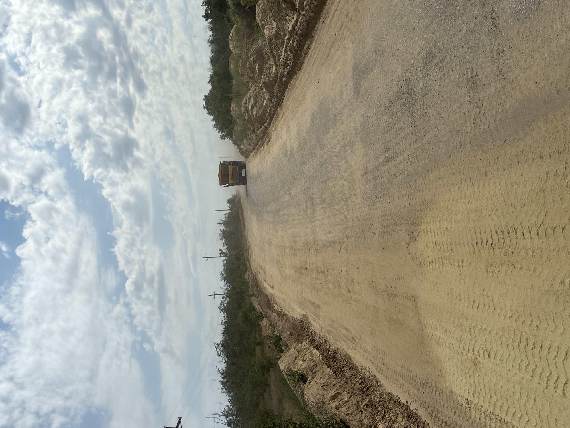 A yellow dump truck is driving on a dusty, unpaved dirt road in a rural or undeveloped area. The truck is kicking up a cloud of dust as it moves away from the camera. The road appears to be under construction or undergoing maintenance, with piles of dirt and sand on the right side of the road. There are some trees and vegetation on the left side of the road and in the background. Power lines and utility poles are visible in the distance. The sky is partly cloudy with a mix of blue sky and white clouds. The overall scene suggests an outdoor, possibly industrial or construction-related environment.