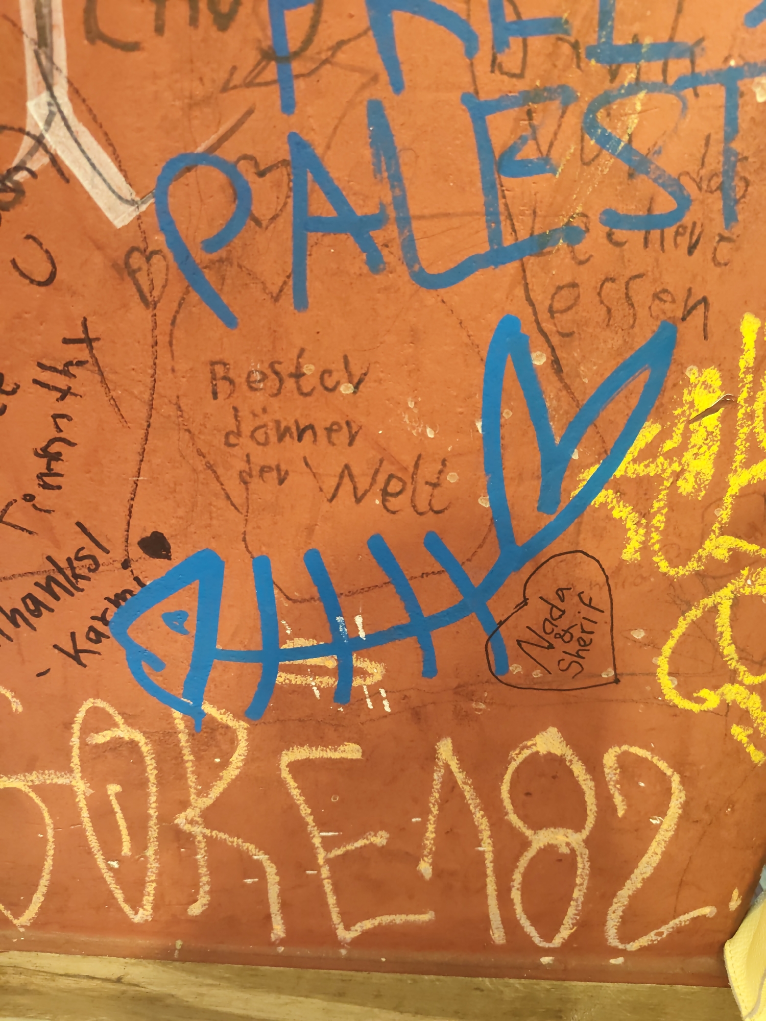 The image displays graffiti on an orange, textured wall, likely in Berlin, Germany, given the presence of German text and a general urban aesthetic.
Dominating the upper portion of the image is large, blue graffiti spelling out "PALEST". Below this, smaller, black graffiti reads "Bestcy döññer der Welt" and "thanks! - Karmi". To the right of "PALEST", faint black scribbles appear to include "believe" and "essen".
In the center, a blue graffiti drawing resembles a fish with a stylized heart. Within the heart, black handwriting states "Nada & Sherif". Yellow chalk graffiti is visible to the right of the blue fish.
At the bottom, large, yellow chalk graffiti spells out "CORE 182". The wall itself shows signs of wear and tear, with some white specks scattered across the surface. The lower edge of the image reveals a glimpse of a light-colored, possibly wooden, surface.