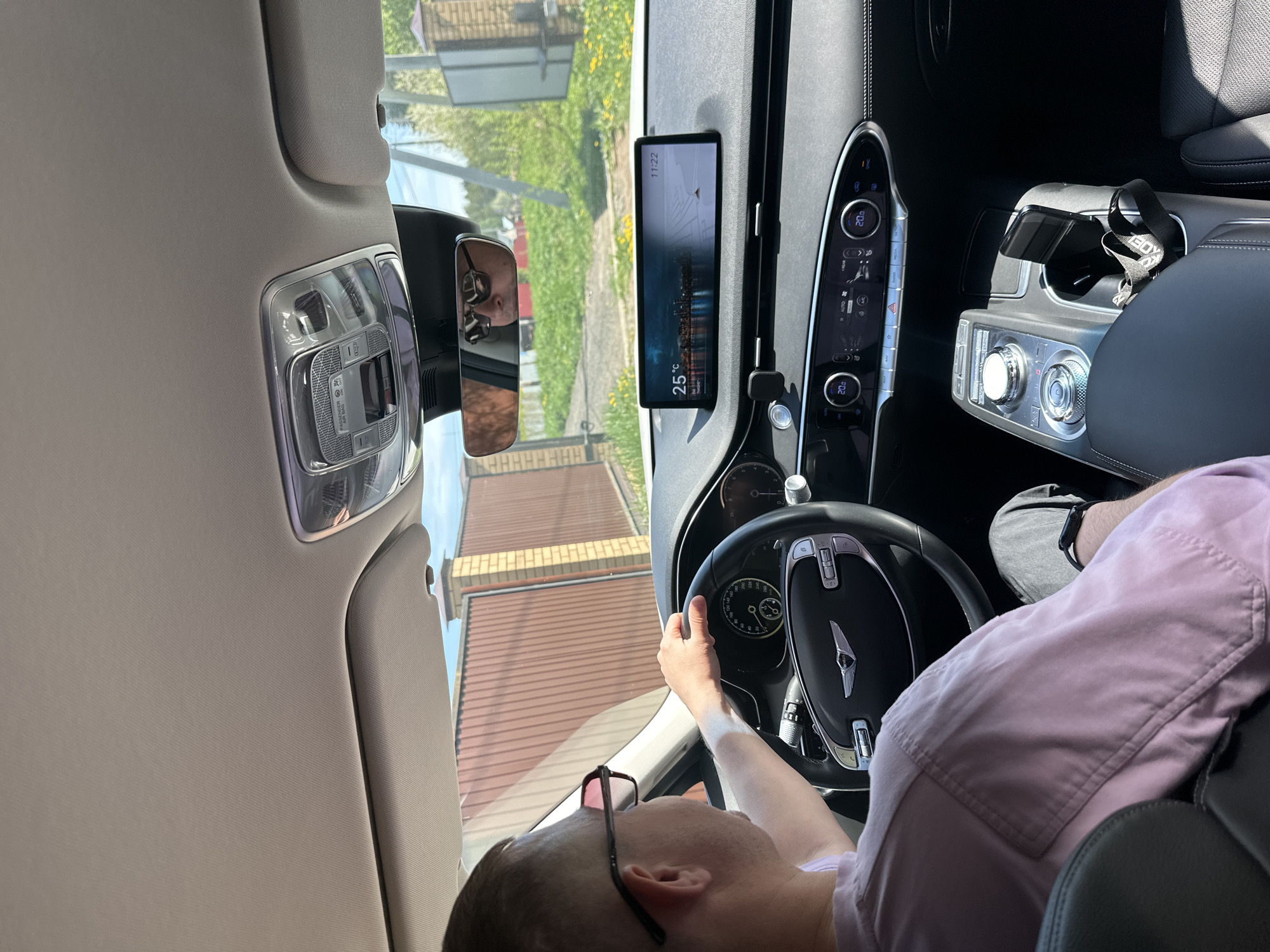 A person is sitting in the driver's seat of a car, possibly in Прикамский, Russia, based on the street visible through the windshield and side window. The person is wearing a pink shirt and glasses, with their hand on the steering wheel. The car's dashboard is visible, including the digital display screen showing a map, the climate control panel, and the gear selector. The rearview mirror shows the reflection of a person's face. The interior lights of the car are also visible. The outside environment includes houses and greenery.