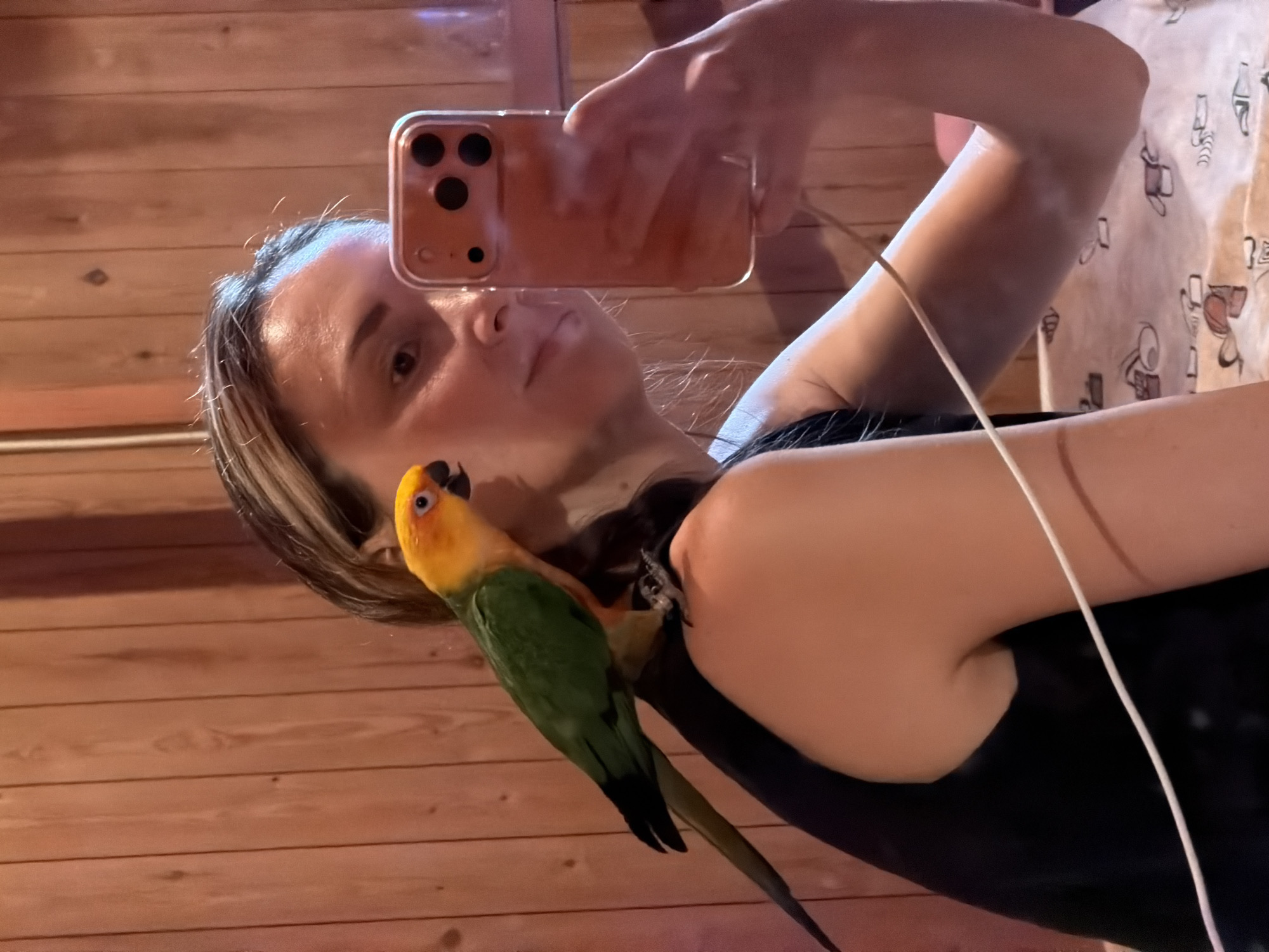 A woman with blonde-streaked brown hair, wearing a black sleeveless top, takes a mirror selfie with her phone. A sun conure parrot is perched on her left shoulder, its green body and bright yellow head visible. The background consists of wood-paneled walls and a patterned bedsheet. The scene takes place indoors.