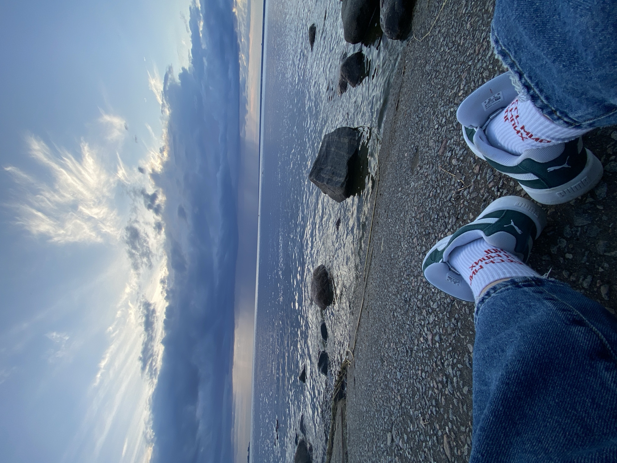 The image shows a person's legs and feet, wearing blue jeans and white socks with red lettering, and green and white sneakers, sitting by the water. The foreground is a rocky shore. In the mid-ground, there are large rocks in the water, with ripples on the surface. The background features a vast body of water stretching to the horizon under a dramatic sky with dark storm clouds and wispy white clouds catching the light. The scene appears to be a waterfront area in Sestroretsk, Russia, possibly during or just before a weather event.