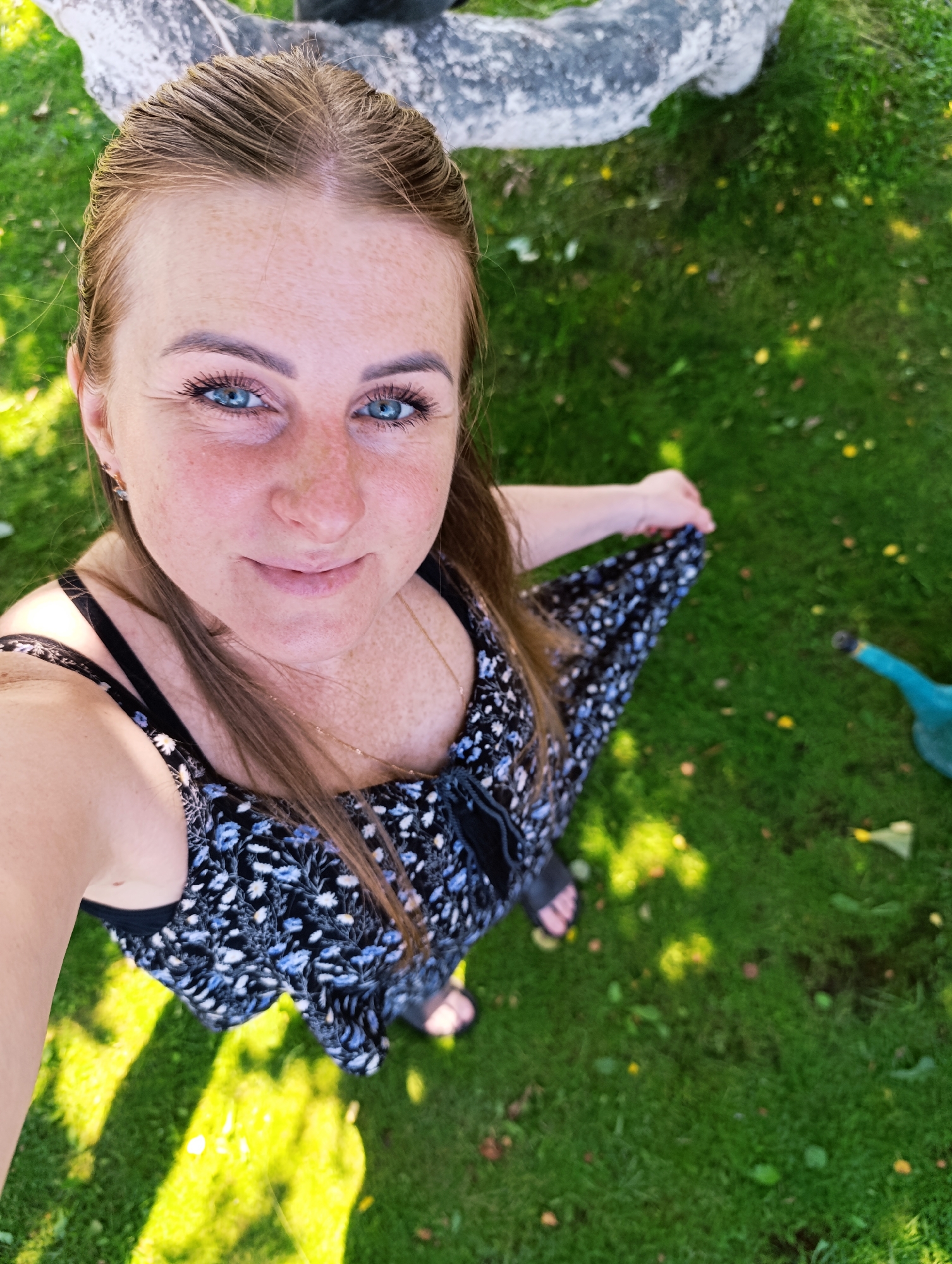 A high-angle, full-body shot captures a woman with light brown hair and blue eyes, wearing a black and white floral dress and black sandals, standing on a grassy lawn. She is looking up and smiling at the camera, holding the hem of her dress with her right hand. Sunlight casts dappled shadows across the grass, and a blue watering can is visible on the right side of the frame. The scene appears to be an outdoor setting, possibly a backyard or park, with a large, light-colored, possibly stone, object in the background.