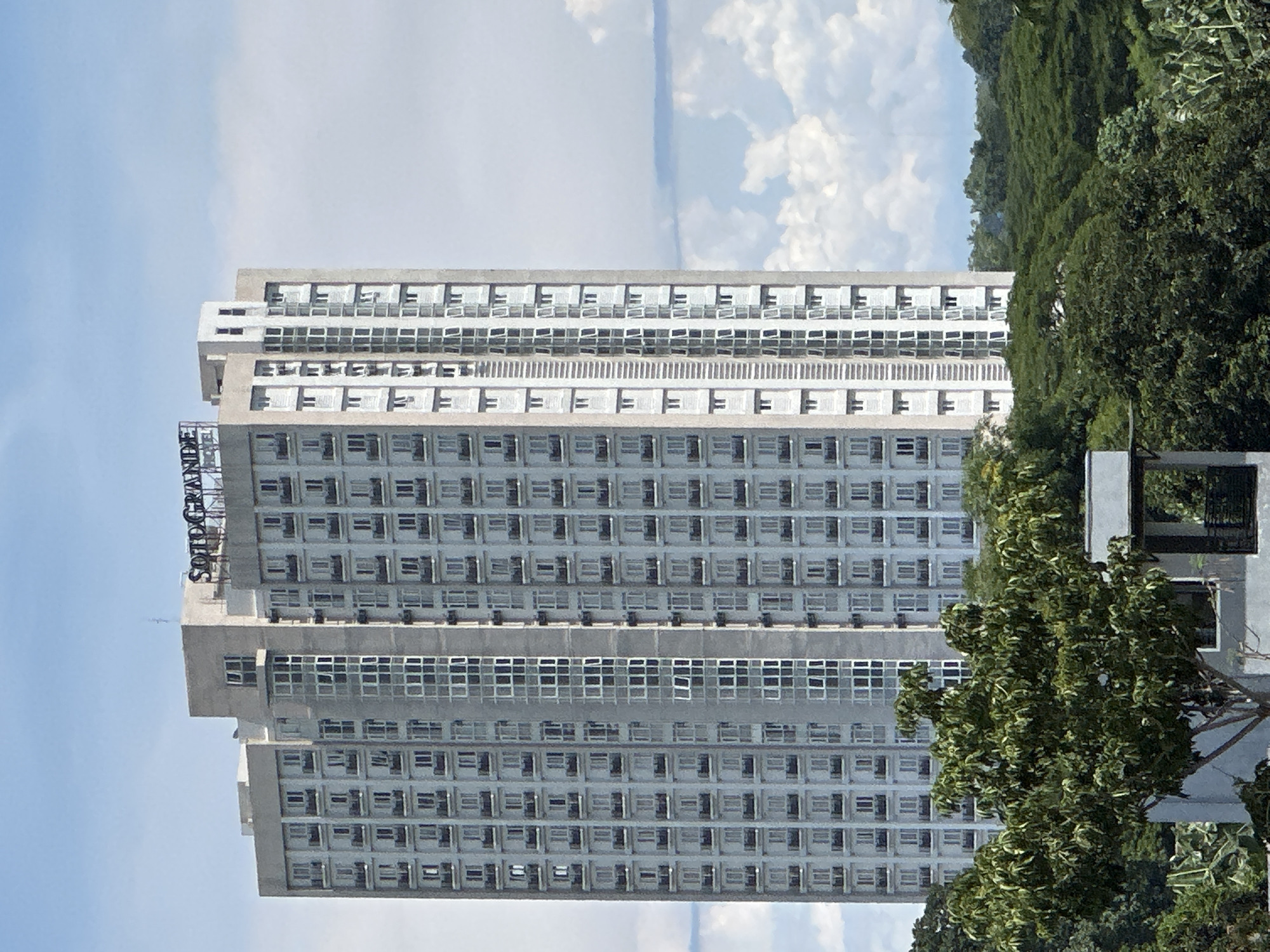 The image depicts the high-rise condominium building "Sotogrande" located in Davao City, Philippines. The building is a multi-story rectangular structure with numerous windows arranged in a grid-like pattern. The sky is visible behind the building, with white clouds. Lush green trees are seen on the right side of the frame, partially obscuring the building's lower section. A structure, possibly a building entrance or support structure, is visible in the lower right corner, partially obscured by the trees.