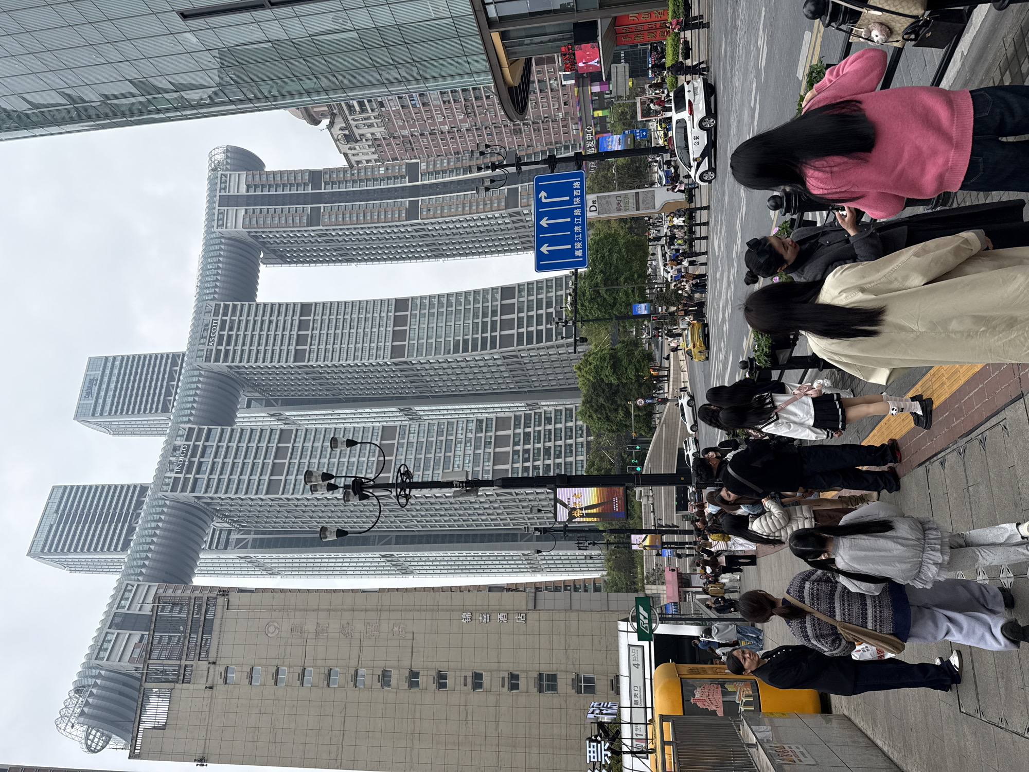 The image shows a street scene in Yuzhong District, China. A group of people stand on the sidewalk, looking towards a set of tall, connected buildings. A street sign in Chinese indicates directions. Several cars and a taxi are visible on the street. Buildings, including Jinxi Hotel, are visible in the background.