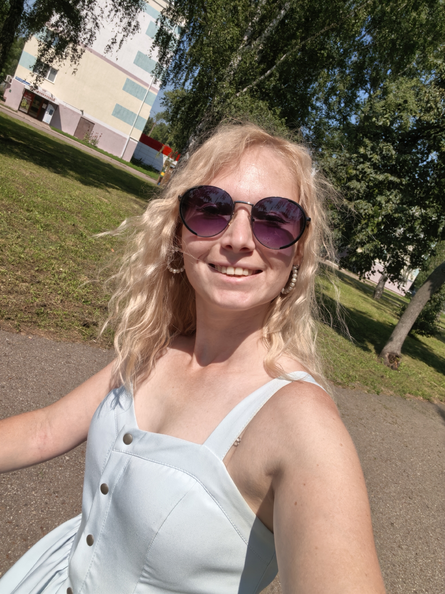 A young adult Caucasian woman with blonde, curly hair and fair skin is taking a selfie outdoors. She is wearing large, round, dark-lensed sunglasses and pearl earrings. Her facial expression is a wide smile. She is wearing a light blue dress with four prominent buttons down the front. The background shows a grassy area, trees, a pathway, and a building with a light-colored facade featuring colored rectangular accents. The scene suggests it is a sunny day. This image was taken in Sterlitamak, Russia.