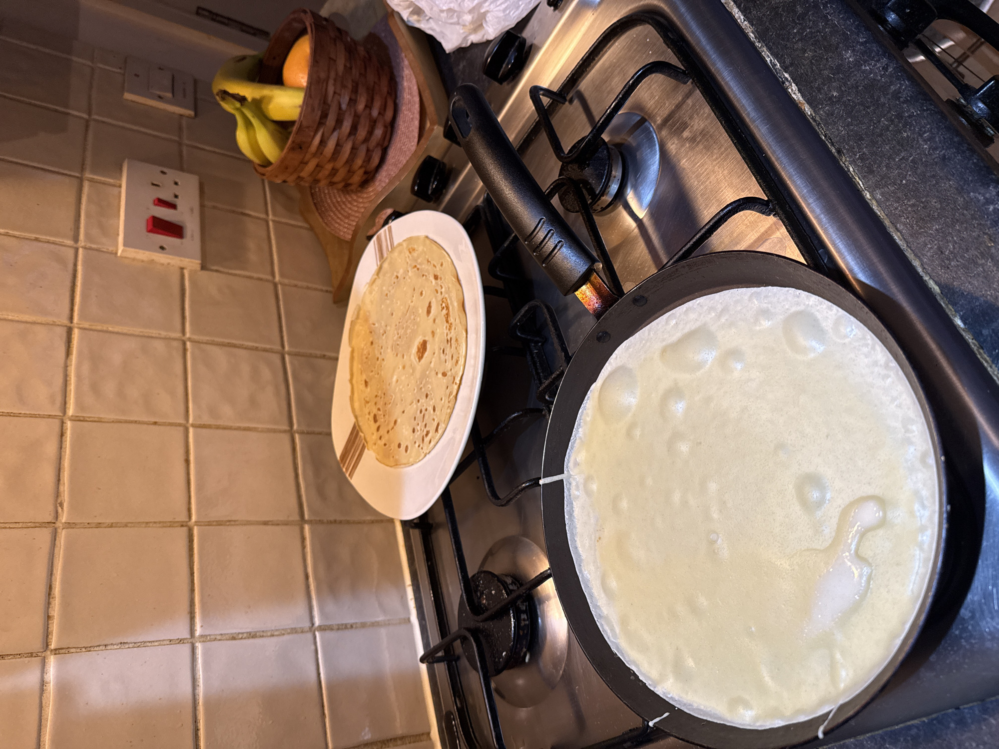 A kitchen scene showing a stovetop with two gas burners. On one burner is a black frying pan containing a freshly poured crêpe, its batter still liquid. On the stovetop next to it, a white plate with brown stripes holds a cooked crêpe. In the background, near a light switch and a double socket, is a wicker basket containing bananas and an orange. The stovetop is set into a black countertop. The wall is covered in beige tiles. The scene is likely domestic. There is no evidence in the image to confirm the location in Great Yarmouth.