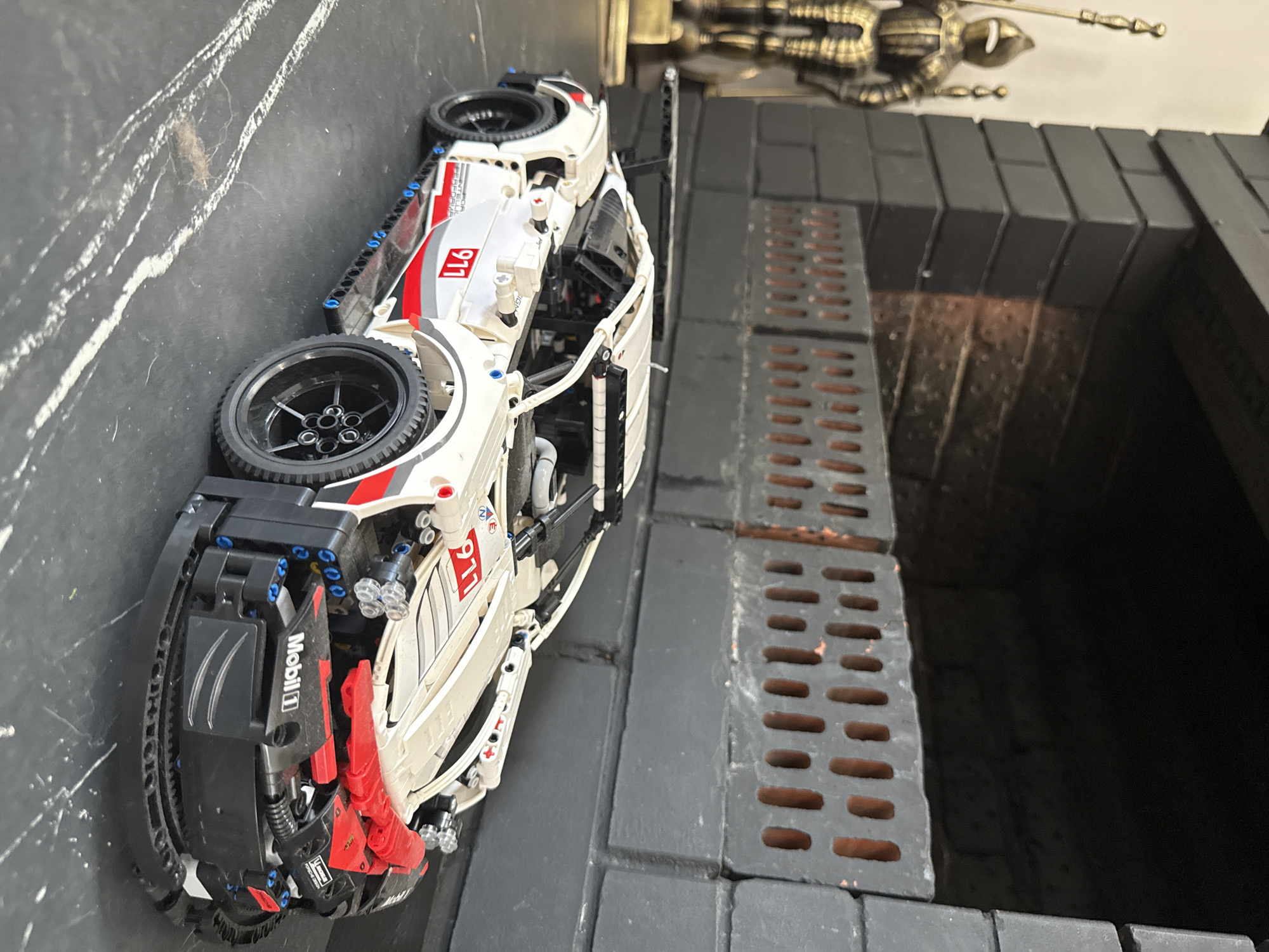 In Хохряки, Russia, a LEGO Technic model of a white Porsche 911 race car with red and black accents is displayed on a dark surface, possibly a table. The car features a prominent rear spoiler, detailed wheels, and racing decals including "911" and "Mobil 1". To the left of the car, a vintage-style metal figurine of a knight in armor holding a spear stands against a dark brick fireplace. The fireplace itself is constructed from dark grey bricks with an opening at the top and a grate-like structure in front of it, revealing reddish-brown bricks behind. The lighting in the scene is somewhat dim, casting shadows and highlighting the details of the car and the knight.