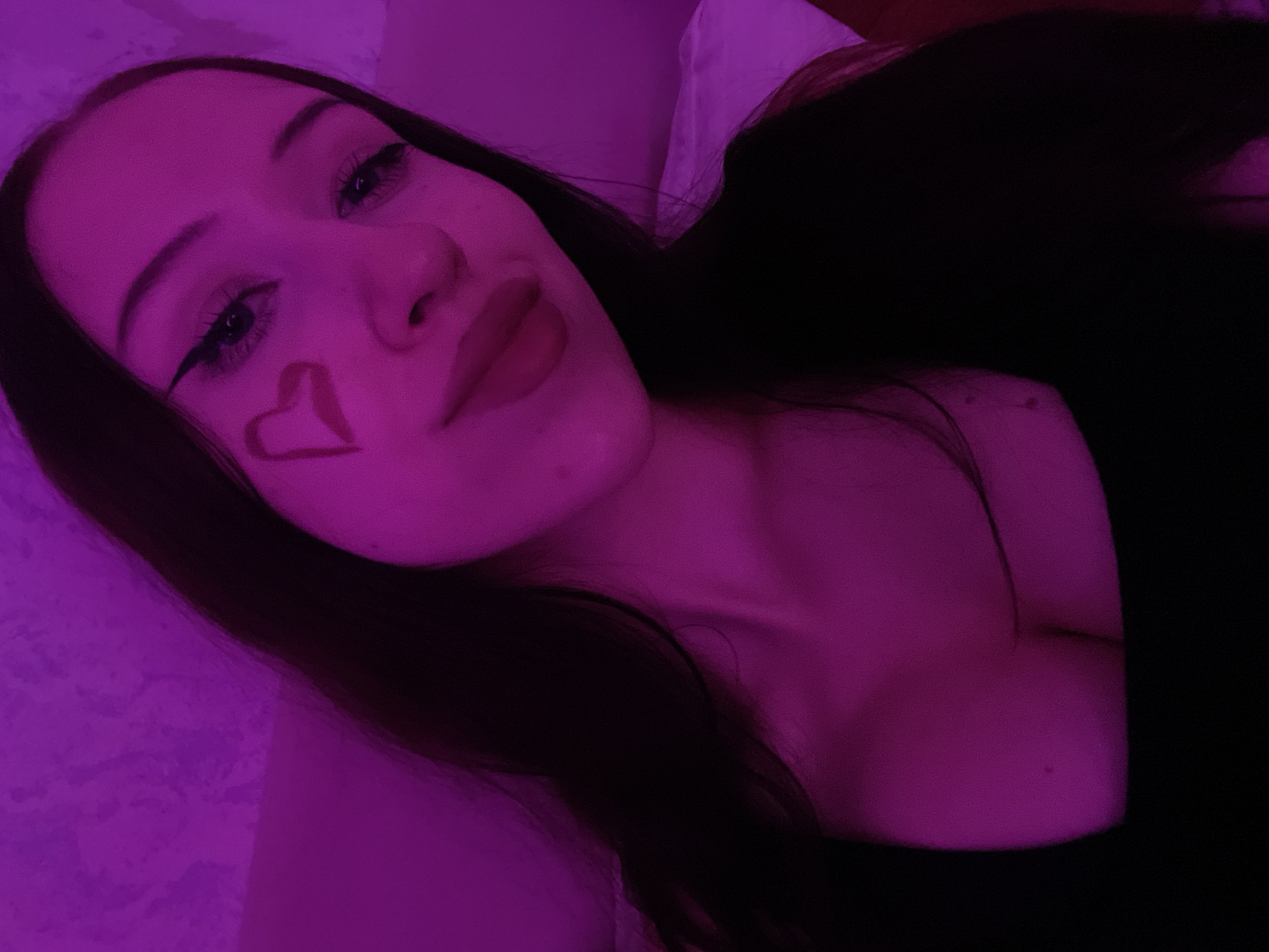 The image is a close-up, selfie-style photograph taken indoors, with a strong pink/purple lighting. The subject is a young woman with long, dark hair. She has makeup on, including winged eyeliner and a red heart drawn on her cheek. She is wearing a dark, low-cut top. The background is dimly lit and appears to be a wall or textured surface. The overall impression is intimate and artistic, likely staged for a personal photograph. There is no indication of the location being Votkinsk, Russia.