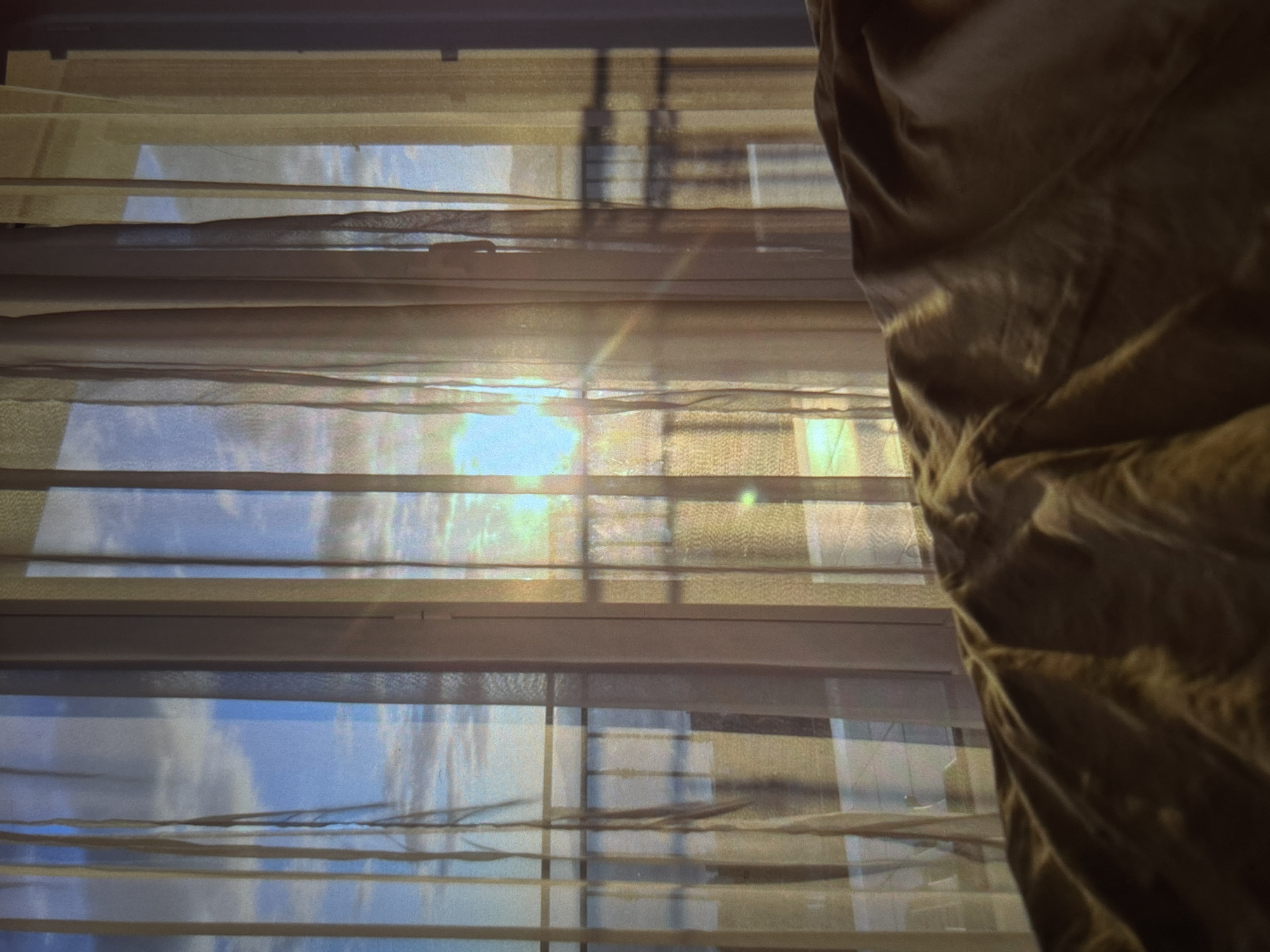 A sunbeam cuts through sheer beige curtains, illuminating the interior of a room. The sun is low in the sky, casting a warm glow that reflects off the window panes. Outside, a railing suggests a balcony or patio overlooking a body of water and a distant shoreline. The light also glints off the surface of the water. In the foreground, a crumpled blanket or duvet in a muted brown hue takes up the bottom portion of the frame. The scene appears to be a peaceful morning or evening view from a window, possibly in a residential area of Knezha, Bulgaria.