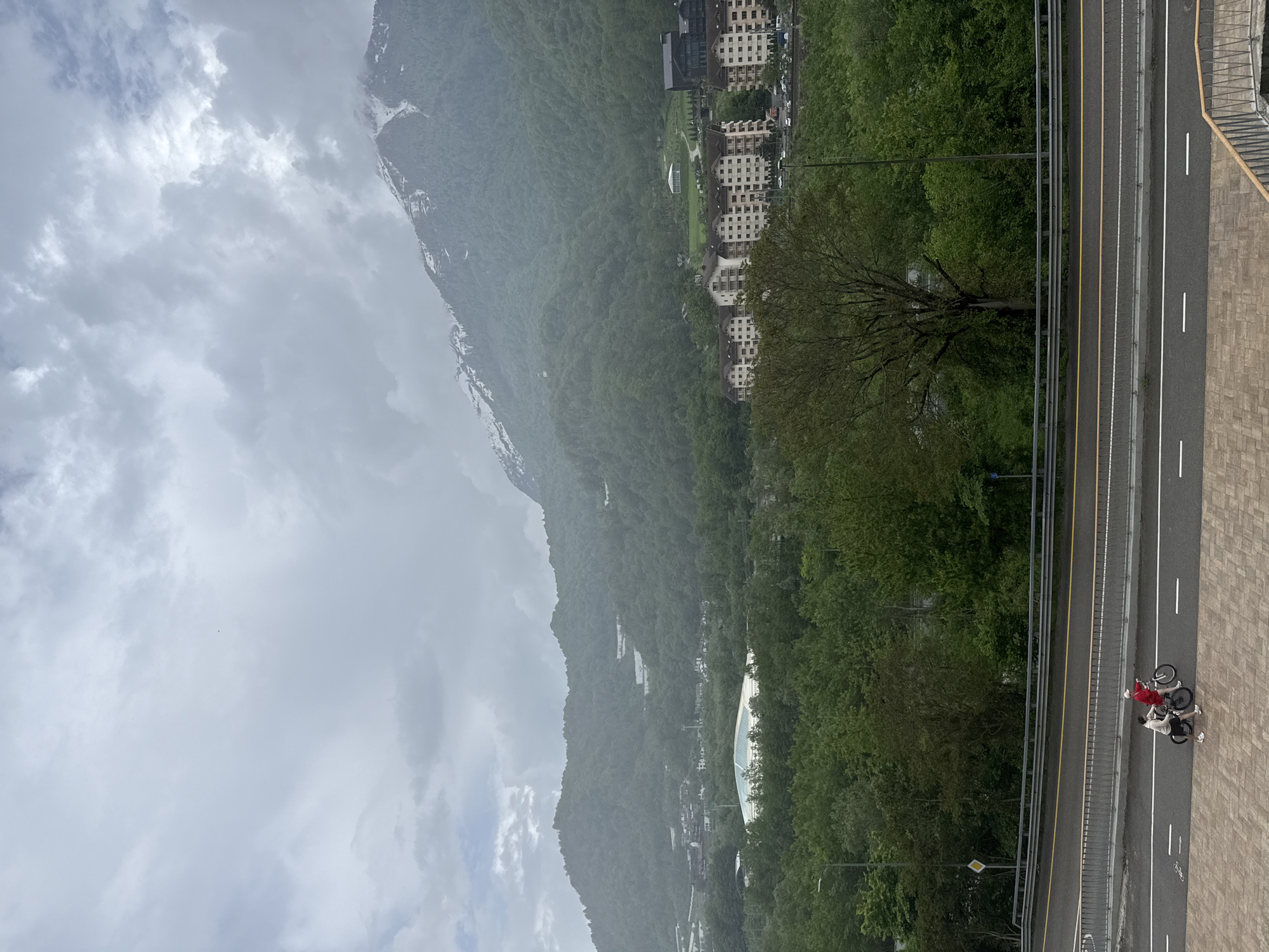 The image shows a mountainous landscape in Krasnaya Polyana, Russia, on a cloudy day. The mountains are covered in dense green forest, with some snow visible at higher elevations. Buildings are nestled among the trees. A highway runs along the right side of the image, with two people standing near bicycles on the road. One person is wearing a red jacket.