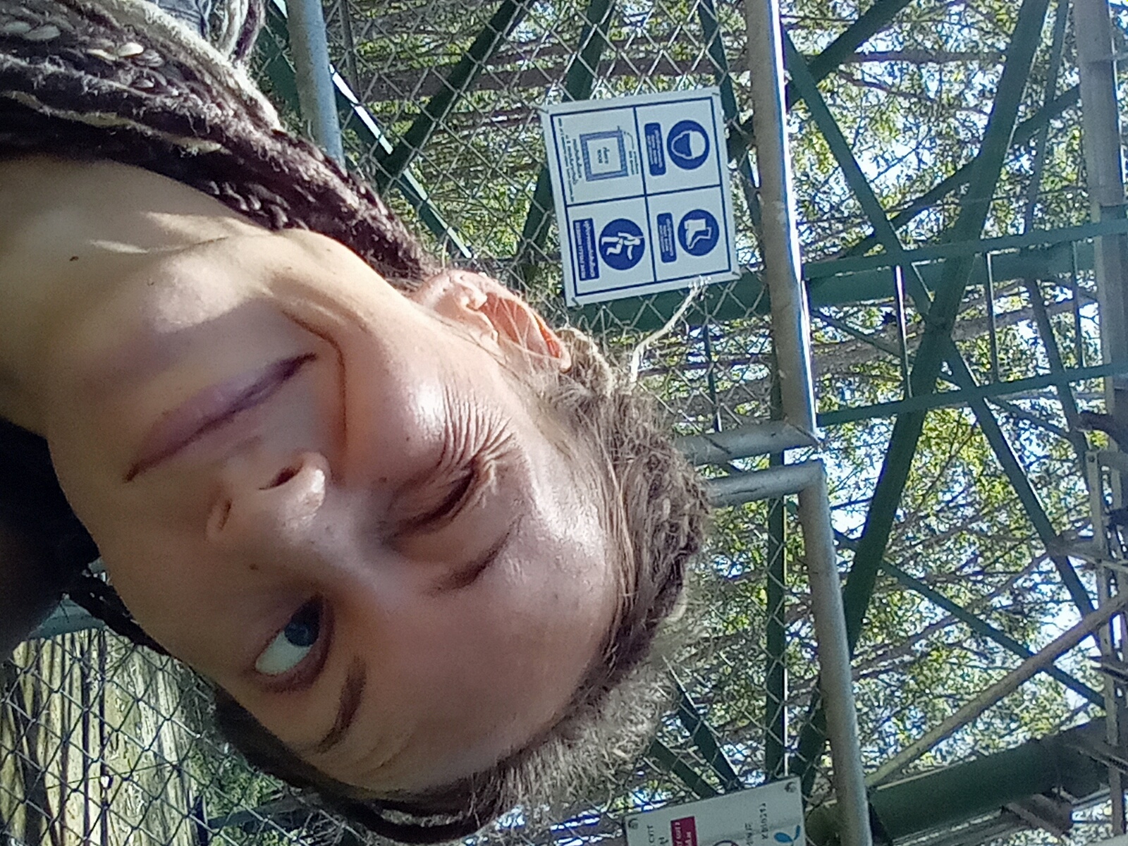 A close-up, upward-angled selfie captures a person with fair to medium complexion and dark dreadlocked hair, winking with their right eye (viewer's left) and displaying a subtle, closed-mouth smile. Their left eye (viewer's right), featuring a blue iris, is open and gazing upward. The background features a chain-link fence with a prominent light gray horizontal metal bar running across it. Attached to the fence on the left is a white square sign containing four blue mandatory action pictograms: a hard hat, safety boots, an illegible textual instruction in Thai, and a figure bending or walking. Partially visible on the right, another vertical sign with Thai text and a blue top suggests "Ko Siboya." Behind the fence and signs, a complex lattice of dark green metal beams forms an architectural structure against a backdrop of dense green trees and patches of blue sky. The scene is outdoors, indicating a bright, sunny day in a natural or protected area, specifically in Ko Siboya, Thailand.