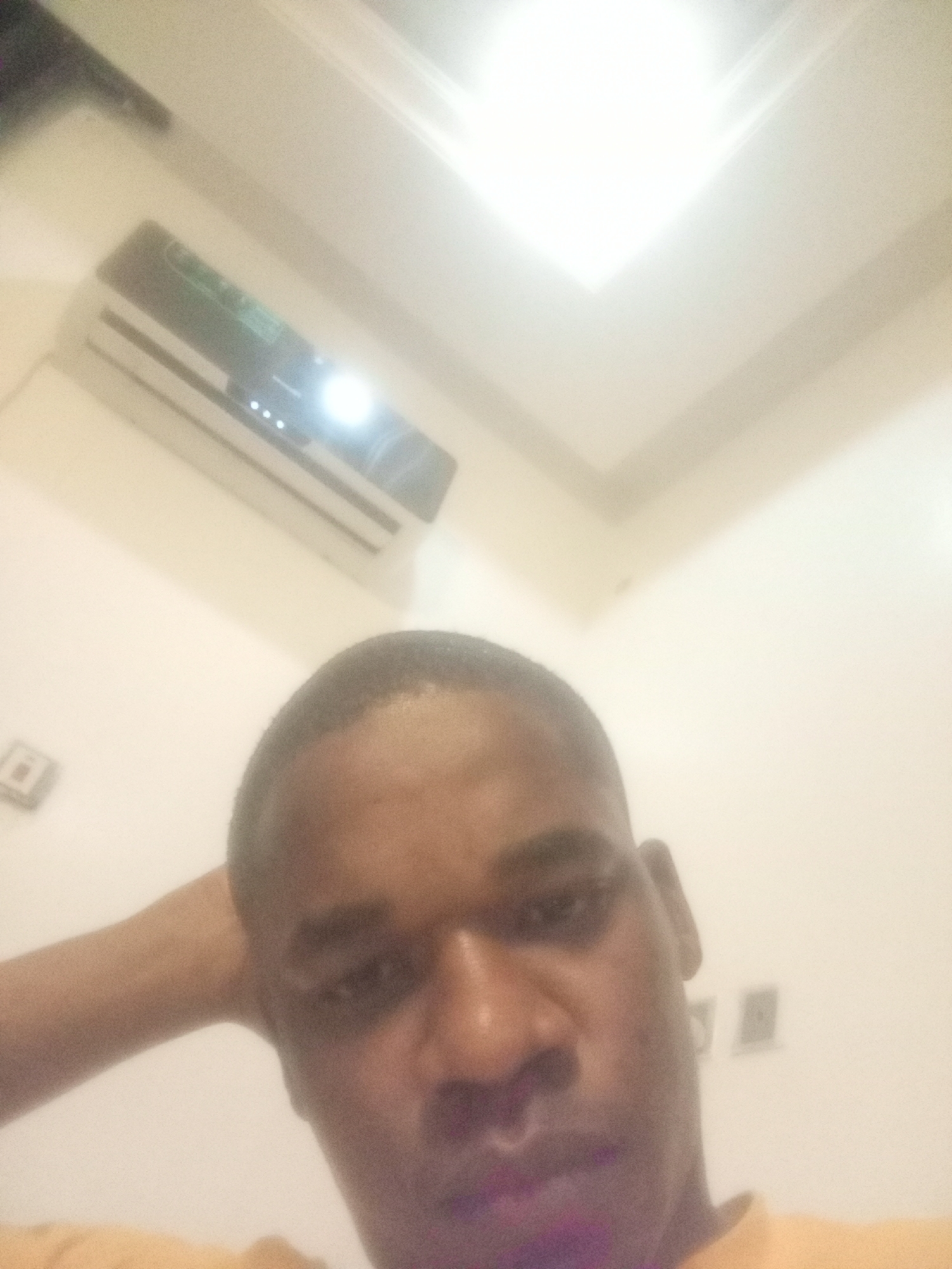 A low-angle, blurred selfie of a dark-skinned person with a short haircut. The person's head is slightly tilted to the right, and their left hand is raised behind their head. The person is wearing an orange garment. In the background is an air conditioner mounted on a wall, a bright rectangular ceiling light, and two electrical outlets on the wall. Location is possibly an interior space in Gwarinpa, Nigeria.