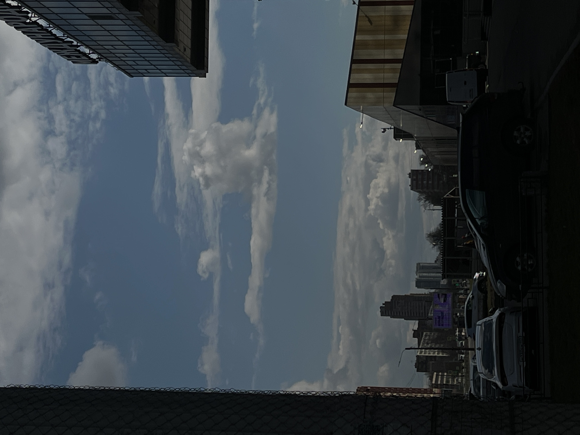 The image shows a view in Saint Petersburg, Russia, with a focus on the sky and buildings. The sky is blue with streaks of white clouds. On the left, the top of a modern glass-clad building is visible. On the right, a different building with horizontal stripes is present, with cars parked along the street. In the distance, the cityscape of Saint Petersburg emerges, with various buildings and structures. A fence is visible in the foreground. The lighting is bright, and the environment seems urban.