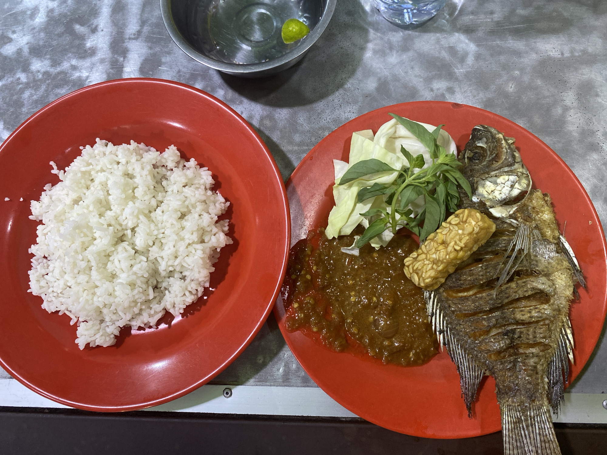 The image shows a meal, likely in North Kuta, Indonesia, given the cuisine. A red plate on the left contains a mound of white rice. Adjacent to it, also on a red plate, is a fried fish, a piece of tempeh, raw cabbage, basil, and a chili paste. A metal bowl with water and a lime wedge is visible in the upper portion of the frame, along with a plastic bottle of water. The setting appears to be a table with a gray, textured surface. The composition is from an overhead angle.