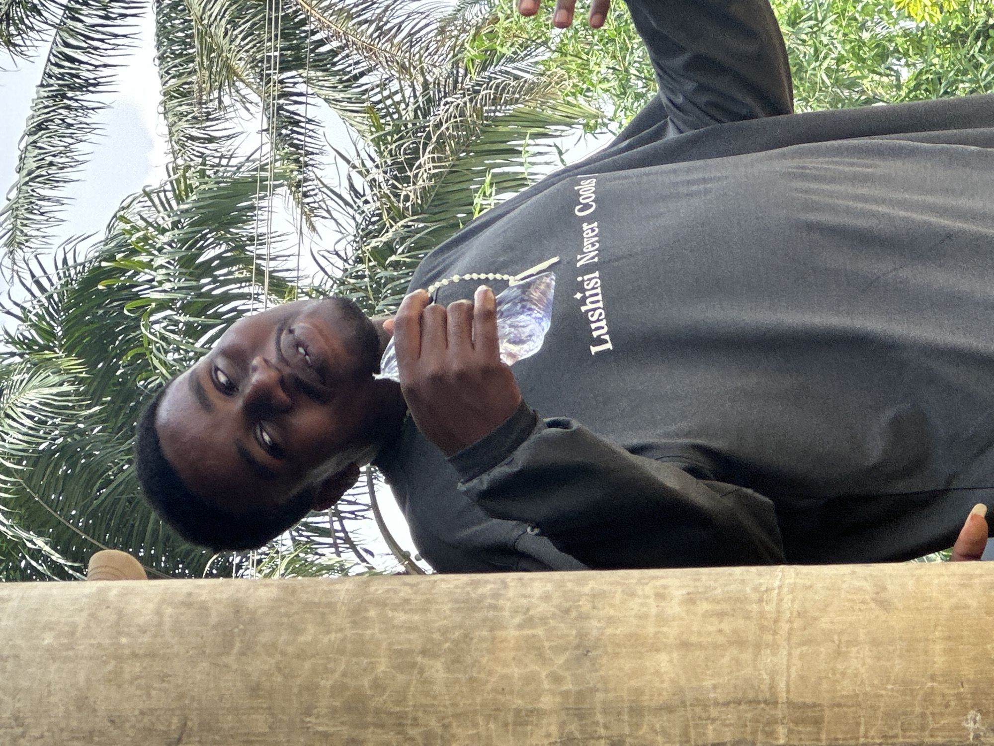 A man stands behind a long, horizontal, light brown wooden surface in Ikot Ekpene, Nigeria. He wears a dark-colored long-sleeved shirt with the words "Lushisi Never Cools" printed on it. He's holding a water bottle in his hand. Lush green palm trees are visible in the background. The sky is partially visible behind the trees.