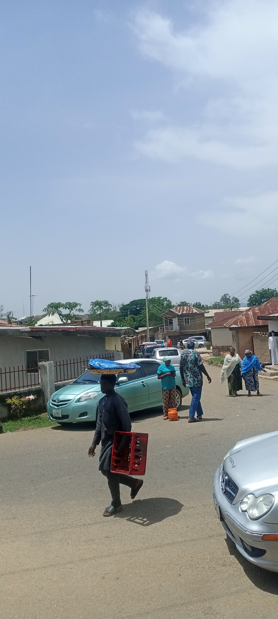 The image shows a street scene in Jos, Nigeria, under a bright sky with scattered clouds. A man in a dark, possibly black, long-sleeved shirt and dark pants is walking on the left side of the street, carrying a red crate in his left hand and a tray covered with a blue cloth on his head. He is wearing sandals.
Several other people are walking further down the street. One woman in a teal shirt and patterned pants is standing near a light blue car with a small orange propane tank next to her. A man in a blue patterned shirt and jeans is walking next to her. Two women wearing head coverings, one in white and one in blue, are walking down the street as well, one using a cane. Several cars are parked along the side of the street.
The background shows houses and buildings with corrugated metal roofs. There is a fenced-in property on the left side of the street. A telecommunications tower can be seen in the distance behind the houses. The asphalt street appears to be dry. A silver car is parked on the right side of the image in the foreground.