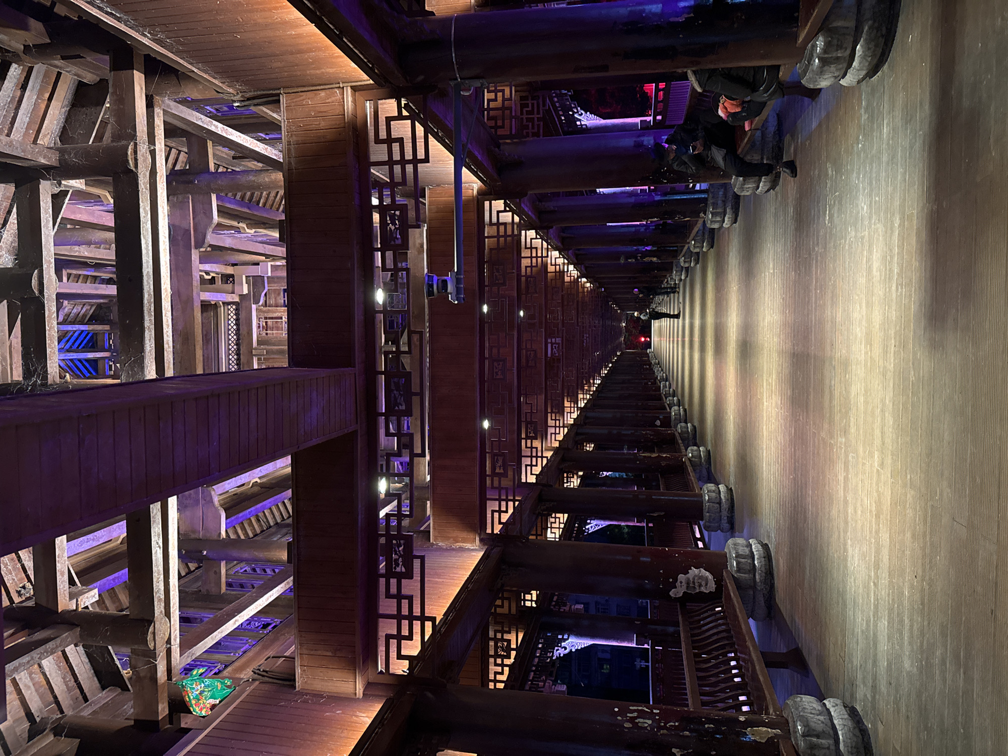 This is an interior view of a traditional wooden structure, possibly a covered walkway or corridor, in Qianjiang, China. The image is taken from a low vantage point, looking lengthwise down the corridor. The structure consists of intricately crafted wooden beams, posts, and lattice-like dividers. The left side reveals the complexity of the framework, with multiple beams intersecting at various angles, some illuminated with purple and blue lights. On the right, people are walking along the corridor. The floor is made of wooden planks, with a series of stone or concrete bases supporting the wooden columns. The distant end of the corridor appears illuminated, drawing the eye towards a vanishing point. Overall, the image highlights the architectural details and construction style of traditional Chinese buildings, with a modern lighting design.