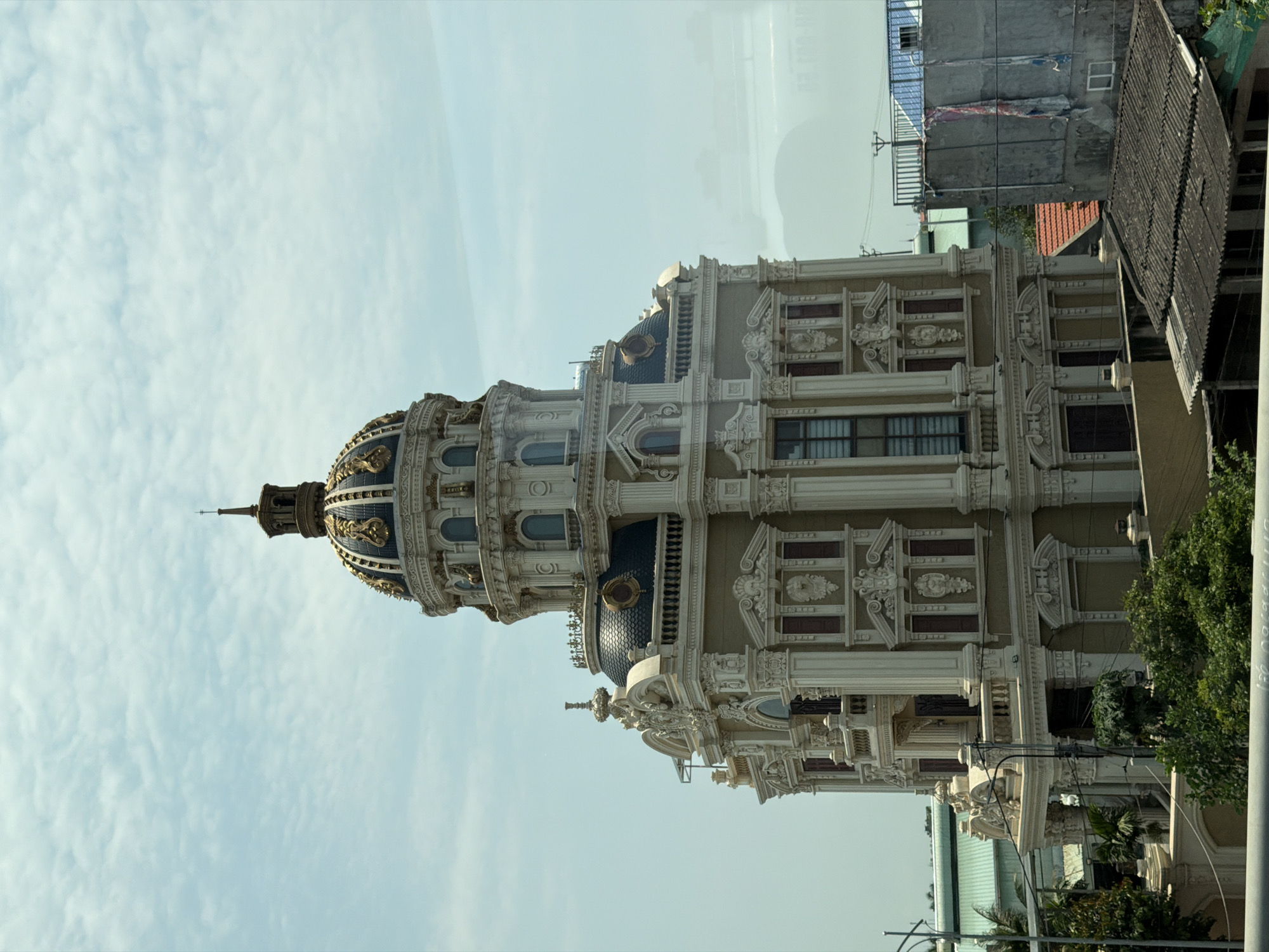 The primary subject of the image is a grand and ornate building, likely located in Hải Phòng, Vietnam, set against a light blue sky with scattered white clouds, indicating daytime. The architectural style suggests a classical or European influence, featuring a prominent central dome topped with a slender spire. The domes are dark blue with intricate gold detailing, adding to the building's luxurious aesthetic.
The facade of the multi-story structure is a light color, possibly cream or beige, and is richly adorned with numerous decorative elements. These include columns, pilasters, elaborate moldings, cornices, and balustrades. Many windows are visible, appearing dark, possibly tinted or reflecting the environment.
To the right and slightly behind the main building, more utilitarian structures are visible, providing a stark contrast in architectural style. One appears to be a concrete building with a blue corrugated roof, while another has a dark, possibly tiled, roof. Green foliage from trees and bushes is interspersed around these buildings, particularly on the lower right side of the frame. No specific activities are depicted, and no people are visible. There is no discernible text within the image.