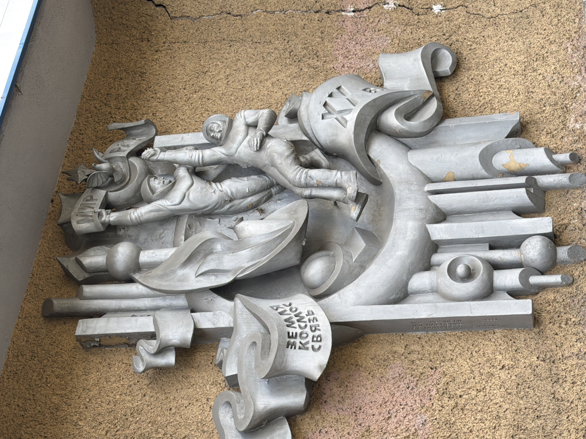 This is a photograph of a relief sculpture mounted on a textured, mustard-yellow exterior wall. The sculpture is made of a light-colored material, likely concrete or plaster, and depicts a scene with two figures and symbolic elements.
The central focus of the sculpture is two male figures in what appear to be stylized space suits, reaching upwards. One figure, on the left, is holding a banner that reads "МИР" (Mir, meaning Peace in Russian). Both figures have helmets, and their bodies are in dynamic poses, suggesting effort or movement. Behind them are abstract, vertical elements resembling pillars or rockets, and a circular shape, perhaps representing a planet or celestial body. To the left of the figures, on a scroll-like element, are inscribed Cyrillic letters that read "ЗЕМЛЯ КОСМОС СВЯЗЬ" (Zemlya Kosmos Svyaz, meaning Earth Space Connection). Below this, a prominent Roman numeral "XXI" is visible on a curved surface.
The wall itself has a rough, stucco-like texture. There is a visible crack running vertically down the right side of the wall, and the paint on the sculpture shows signs of wear and peeling in places, revealing darker undertones. The top of the sculpture is close to a light-colored overhang, possibly the underside of a roof or balcony, with a blue trim visible at the far edge.
This sculpture is located on the exterior of the Zhytomyr State Technological University (Zhytomyr Polytechnic State University) in Zhytomyr, Ukraine. It is a monumental work symbolizing scientific progress, space exploration, and peaceful aspirations.