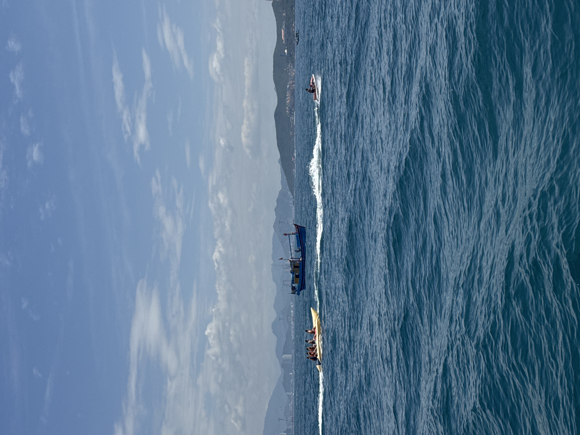 In the city of Nha Trang, Vietnam, a bright blue sky with scattered white clouds stretches over the ocean. The water is a deep blue, with gentle waves rippling across the surface. In the middle distance, a yellow banana boat, carrying five individuals, speeds across the water, leaving a white wake. To the right of the banana boat, a blue and yellow fishing boat sails. Further to the right, a person on a red jet ski is moving at a high speed, creating a significant splash. In the background, a lush, green mountainous coastline is visible, dotted with buildings and structures. The overall scene captures a lively day of water activities in Nha Trang.