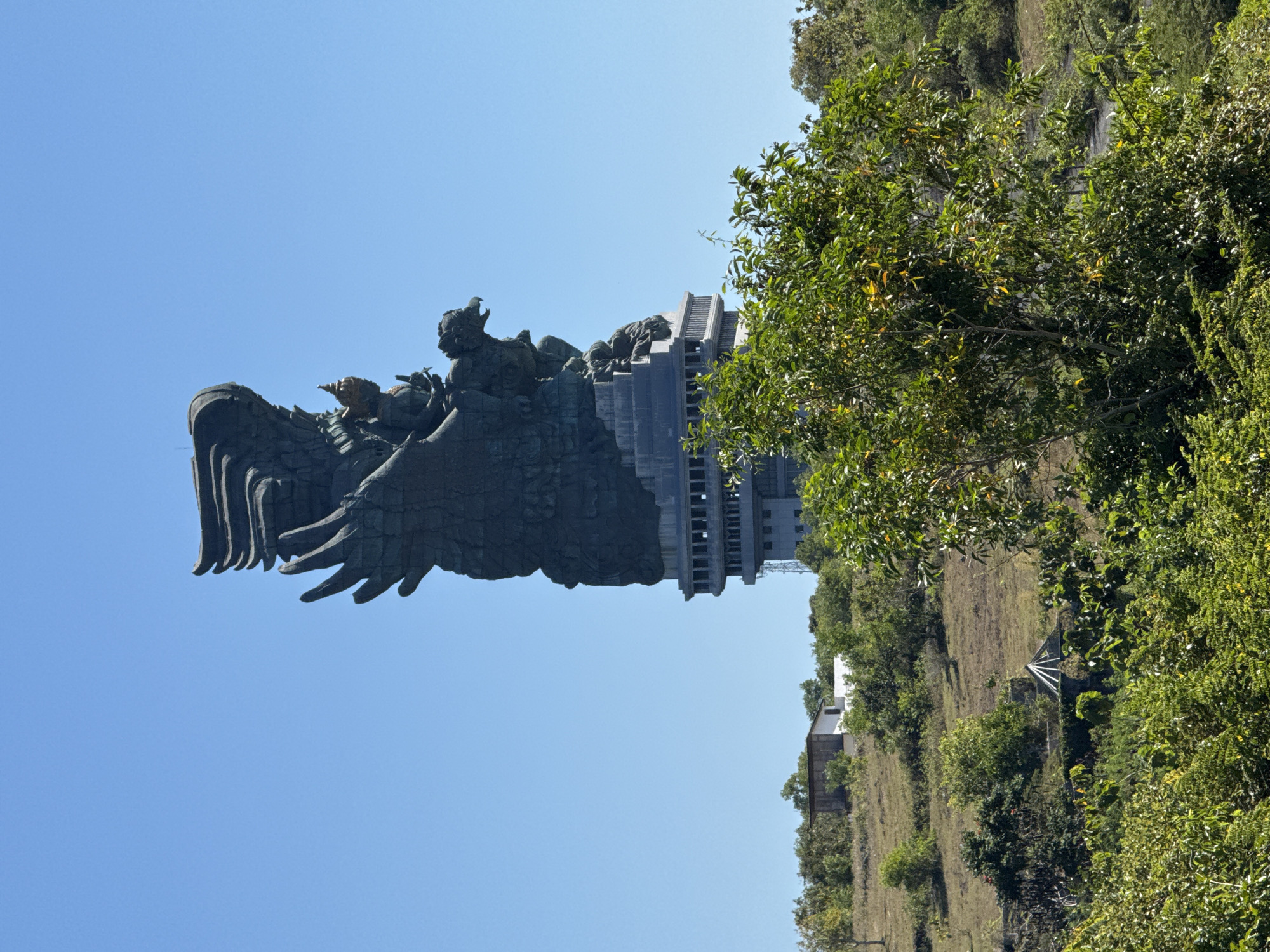 The image shows the Garuda Wisnu Kencana statue in Kuta Selatan, Indonesia. The statue, depicting Lord Vishnu riding the mythical Garuda bird, dominates the left side of the frame. The statue is primarily stone-colored, with some golden accents. The sky is clear and blue. To the right of the statue, a building with many stacked levels is visible. Lush green foliage fills the right portion of the image, obscuring the landscape. In the lower portion of the image, what appears to be a hill is visible. The entire image is rotated 90 degrees counter-clockwise.
