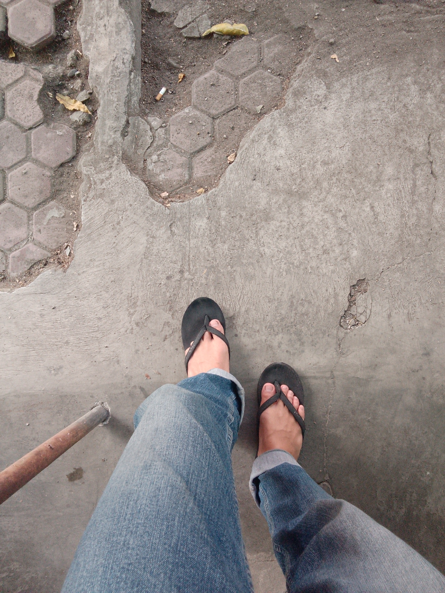 The image is a first-person perspective shot from a standing position, looking down at the ground. The foreground features a person's legs clad in blue denim jeans, with the cuffs rolled up. The person is wearing black flip-flops on both feet. The ground is a mixture of concrete and hexagonal paving stones, with some areas showing wear and tear, including cracks and missing sections. Debris, such as dry leaves and a discarded cigarette butt, are scattered on the ground. A rusty metal pipe extends from the lower left of the frame into the ground. The scene appears to be an outdoor urban environment, possibly a sidewalk or walkway, in Trenggalek, Indonesia.