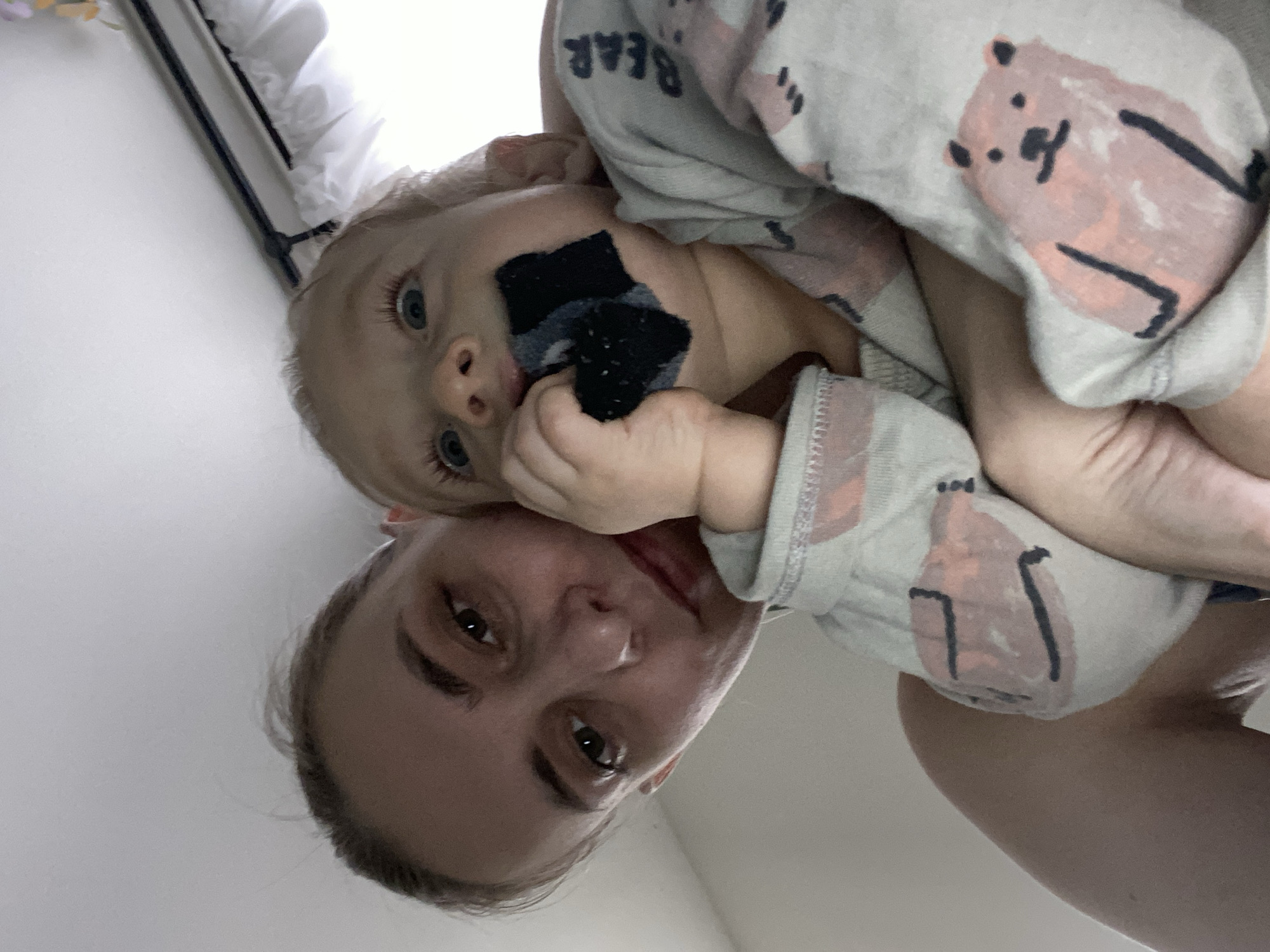 An adult female and an infant are captured in a close-up, upward-angled photograph, indoors in Chaikovsky, Russia. The adult female is positioned on the left, looking directly at the camera with a neutral expression. She has light skin, brown eyes, and her light brown hair is pulled back. Her bare left shoulder is visible. To her right, the infant is held, facing generally towards the camera with wide blue eyes. The infant is wearing a light-colored long-sleeved garment patterned with pink bears and the word "BEAR" in dark blue. The infant's right hand holds a dark, textured fabric object, approximately 4x3 cm and irregular in shape, to its mouth. The background is predominantly a bright, plain white surface. In the upper right corner, a dark curtain rod, white ruffled curtains, and a faint floral decoration are partially visible, suggesting a window as a light source.