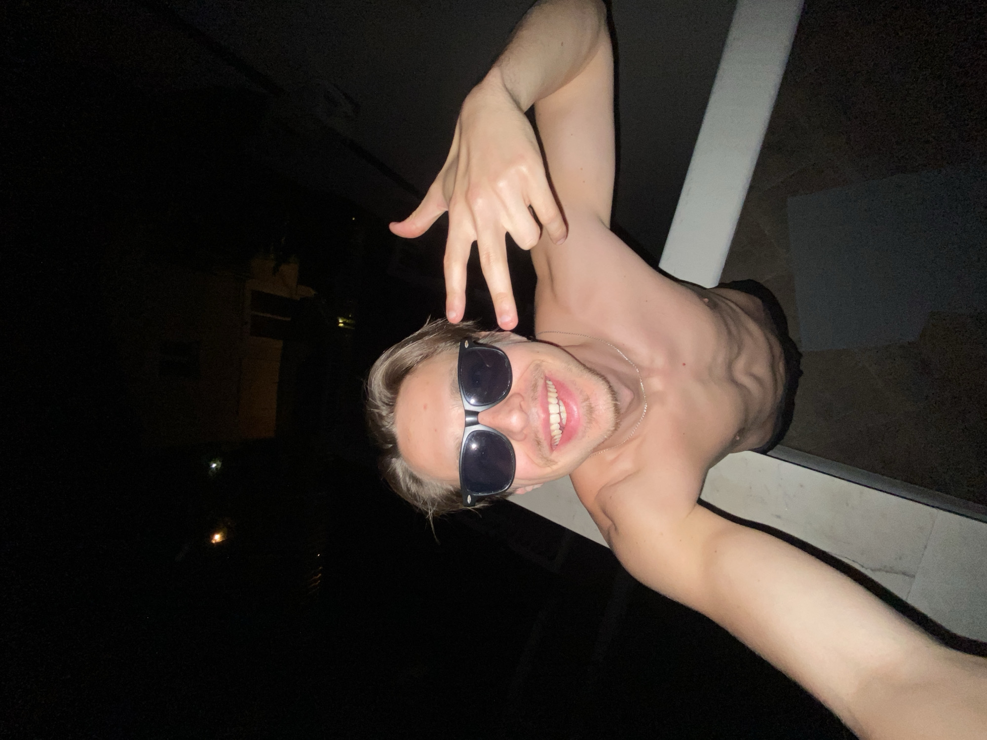 A shirtless man with blonde hair and sunglasses is leaning over a white railing at night. He is smiling and making a hand gesture. The background is dark, but there are some lights visible in the distance. The location is Šušanj, Montenegro.