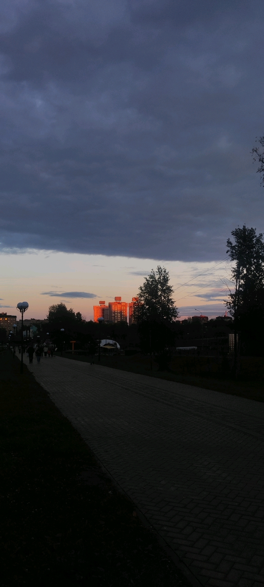 A vertical, low-angle shot captures a city park path at dusk. The path, made of interlocking gray bricks, leads into the foreground from the right, with grass and dark trees lining its left side. Several indistinct figures walk along the path. In the background, a cluster of tall, red-orange buildings is illuminated by the setting sun, contrasting with the dark, heavy clouds overhead. Trees and power lines are silhouetted against the twilight sky. The scene is likely the Victory Park in Cheboksary, Russia, known for its paths and recreational areas.