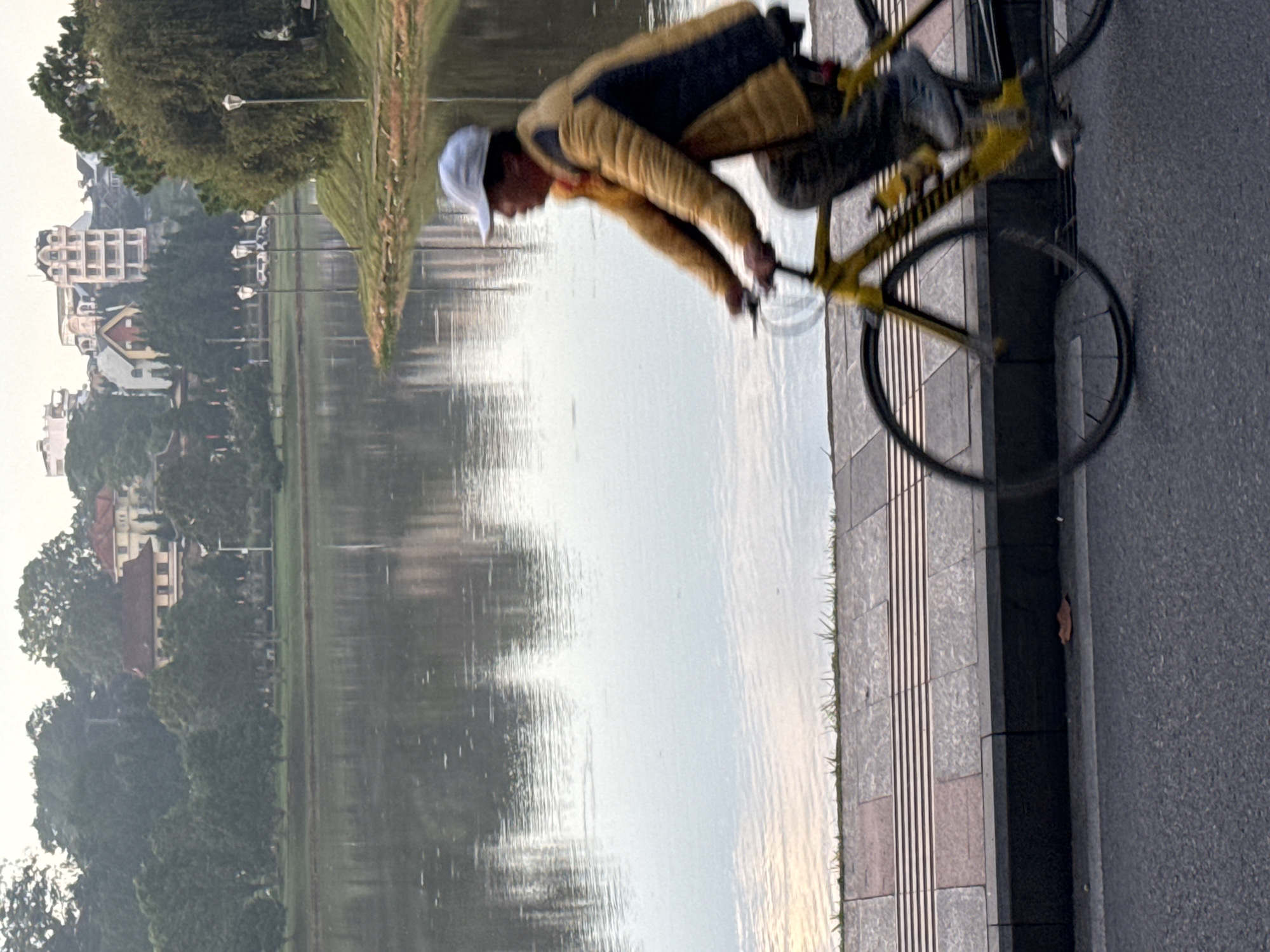 A person is cycling on a paved path next to a calm body of water. The cyclist is wearing a light-colored baseball cap and a yellow jacket. The path has a textured surface with gray and pinkish-brown tiles, and a dark gray curb. Beyond the path, the water reflects the sky and trees. In the background, across the water, are buildings and lush green trees. The scene appears to be at Xuan Huong Lake in Da Lat, Vietnam, given the characteristic architecture and the presence of a lake with surrounding parkland.