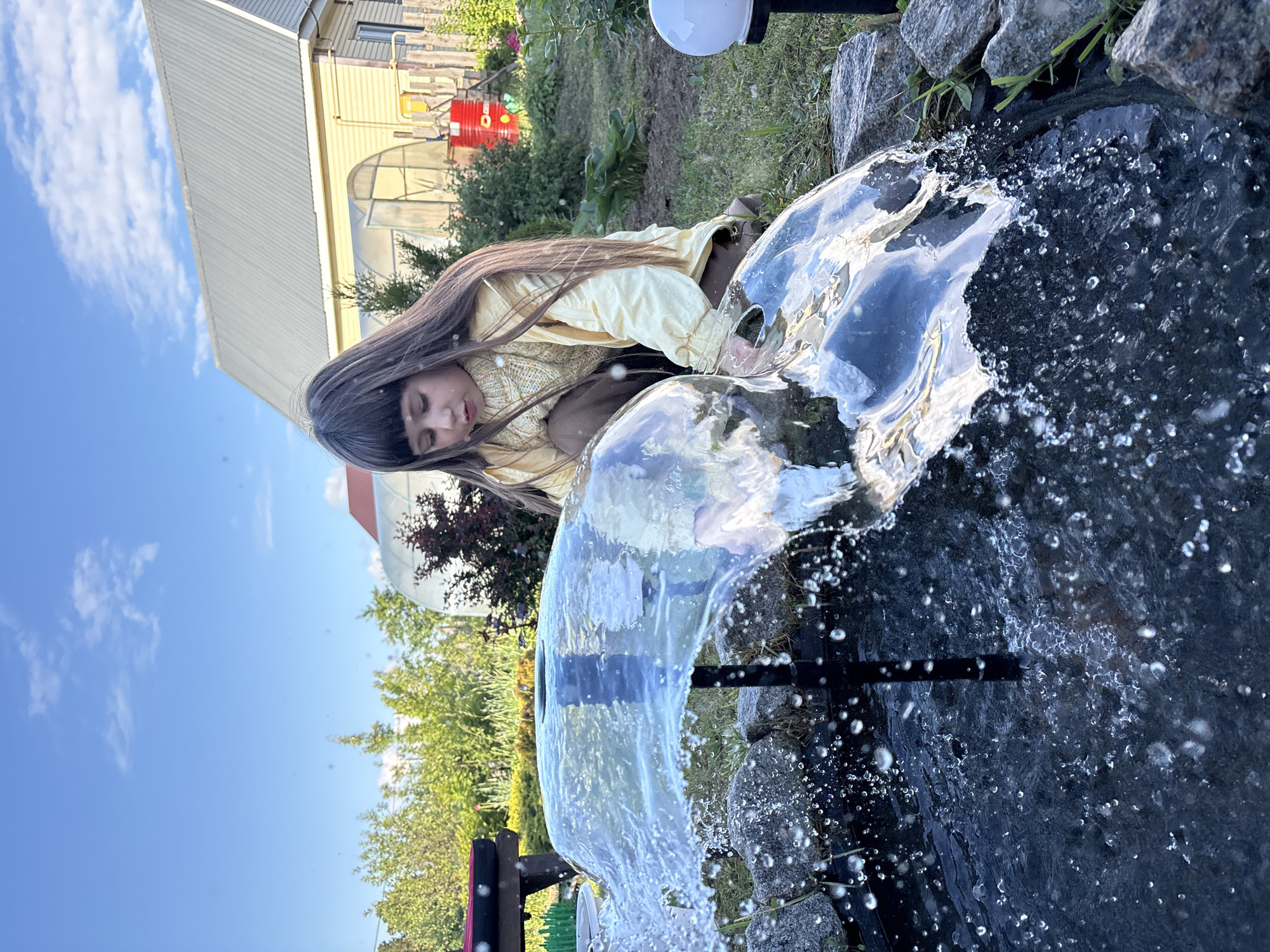A young person with long, dark brown hair is depicted in an outdoor garden setting, leaning over a dark-colored water feature. The image is presented in a landscape orientation but is rotated 90 degrees clockwise, causing the sky to appear on the left side.
The person, dressed in a light yellow jacket over a textured, light-colored top and dark pants, is crouched or kneeling, closely observing the actively splashing and flowing water. Her eyes are cast downwards, conveying a sense of peaceful contemplation or focus on the water. The water in the feature is clear, creating visible ripples and numerous droplets as it moves. Several grey rocks surround the edge of the pond, and a black pipe or structure is partially submerged within the water.
In the background, under a bright blue sky dotted with scattered white clouds, there is a light-yellow building featuring a large corrugated metal roof and an attached greenhouse-like structure with a rounded top. Green vegetation, including trees, bushes, and grass, frames the scene. Other background elements include a red barrel or tank and a white outdoor light fixture on a black pole. The scene takes place during a clear, sunny daytime, likely in a garden or residential area in Chaikovsky, Russia. No visible text is present in the image.