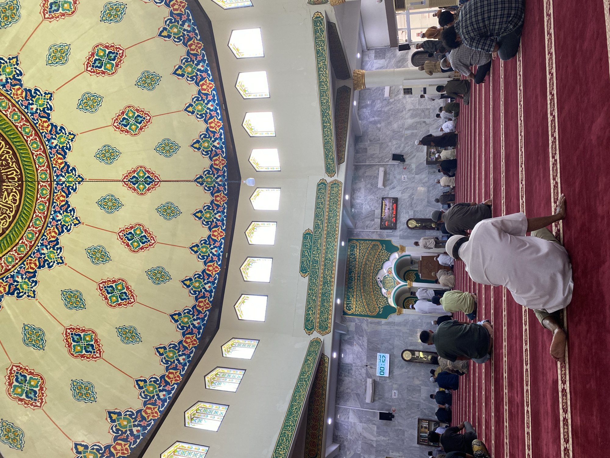 The image depicts the interior of a mosque in Lhokseumawe, Indonesia, filled with approximately 30 individuals, predominantly men, seated on a red carpeted floor arranged in rows facing the front. The mosque features a domed ceiling adorned with intricate, colorful geometric patterns and Arabic calligraphy. Natural light filters through arched windows high on the walls. Various decorative elements, including green banners with golden Arabic script and architectural details like pillars and arched entrances, are visible. Several individuals are engaged in prayer or quiet reflection, while others are observing. Digital clocks and electronic displays are mounted on the walls. The overall atmosphere suggests a religious gathering or service.