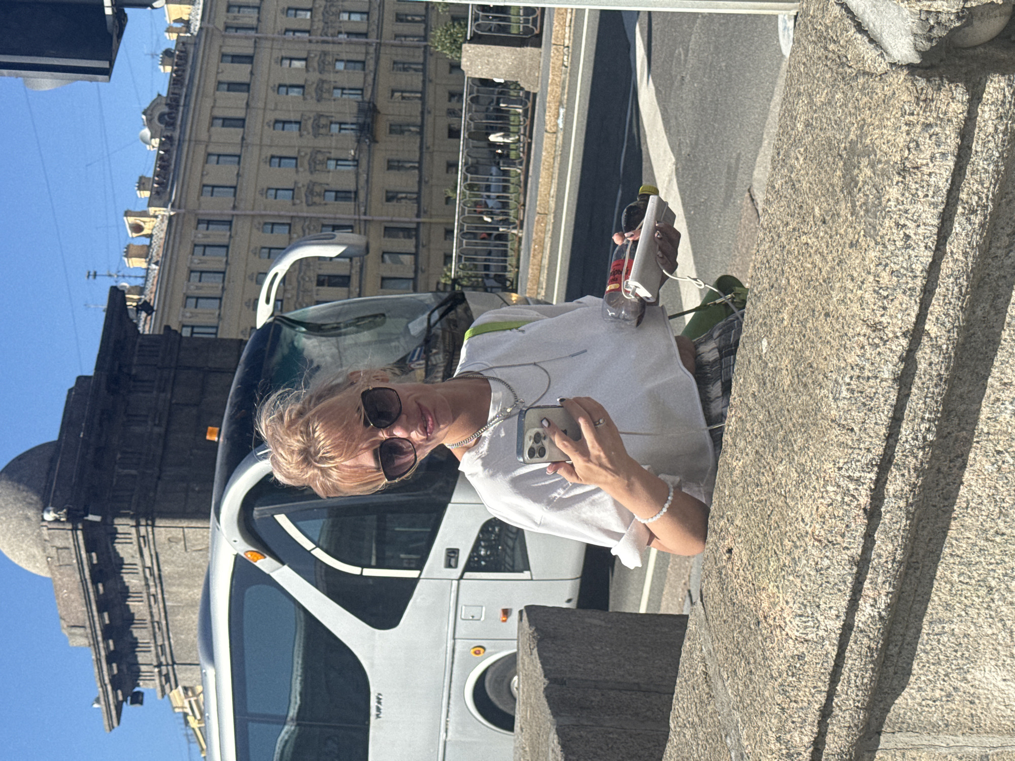 A woman with blonde hair, wearing sunglasses, a white t-shirt, and a bracelet, takes a selfie in front of a white bus. She is holding a smartphone and a plastic bottle with a yellow cap. Behind her, a large, ornate building with many windows stands under a clear blue sky. The scene appears to be on a sunny day, possibly in the afternoon, given the bright lighting and shadows. The bus seems to be a tour bus, possibly in a tourist area of Saint Petersburg, Russia, given the architectural style of the buildings. The woman's expression is relaxed and she appears to be enjoying her outing. The image is rotated, with the ground appearing on the right side of the frame and the sky on the left. There is some visible text on the bus, but it is too blurry to read.