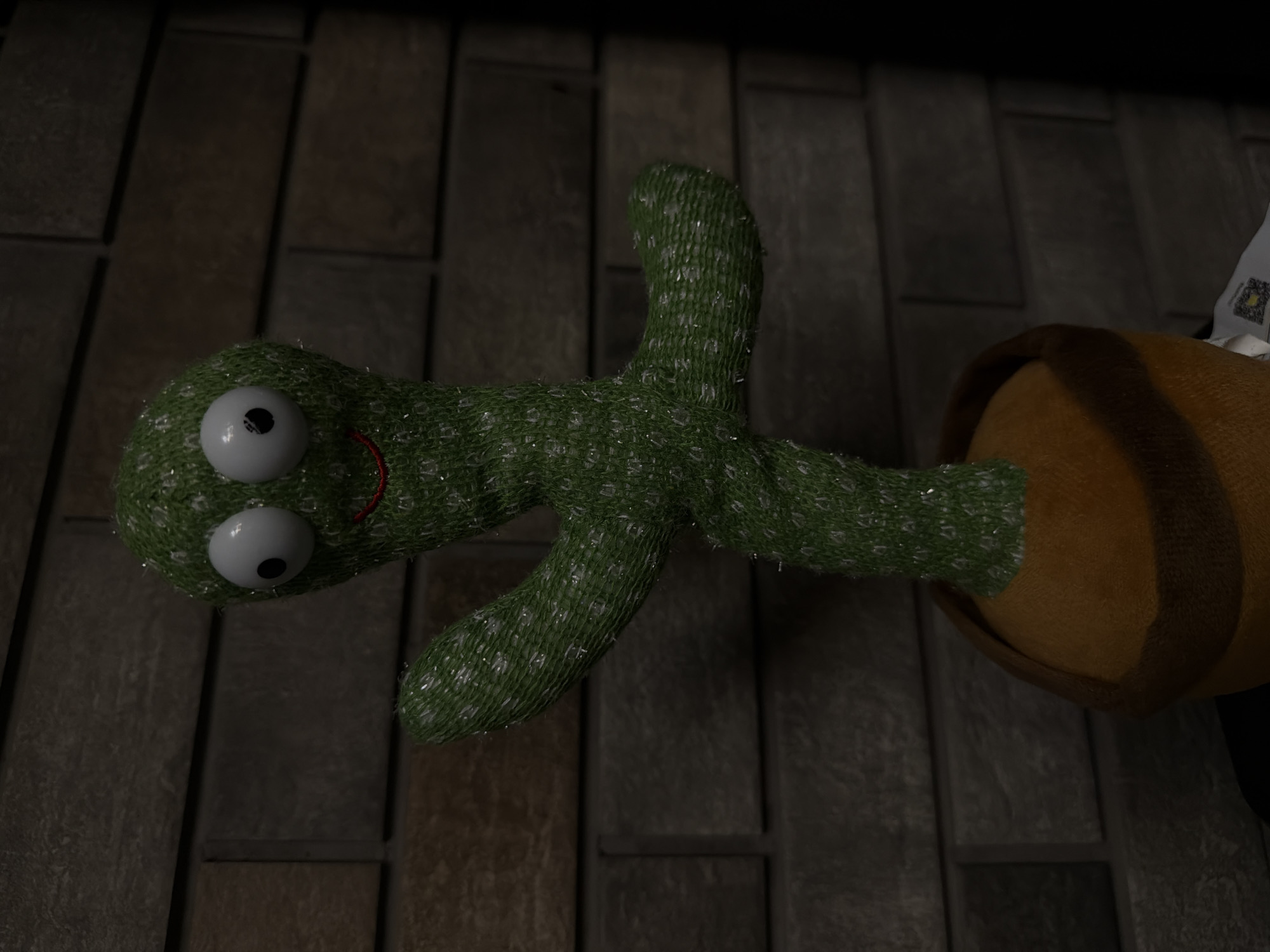 A plush, green cactus toy with large, round, white eyes and a red, smiling mouth stands in a brown pot against a dark brick wall. The toy has two arms extended outwards, one pointing upwards and the other downwards. The cactus is textured and appears to be made of knitted material with shimmering threads. The background consists of horizontal and vertical brickwork, with some shadowing and discoloration, suggesting it is an indoor environment. A small white tag with a QR code is partially visible at the bottom right of the image. The scene is lit from the front, casting shadows on the brick wall. There are no people present in the image. The location of this image in Bila Tserkva, Ukraine is not discernible from the visual information provided.