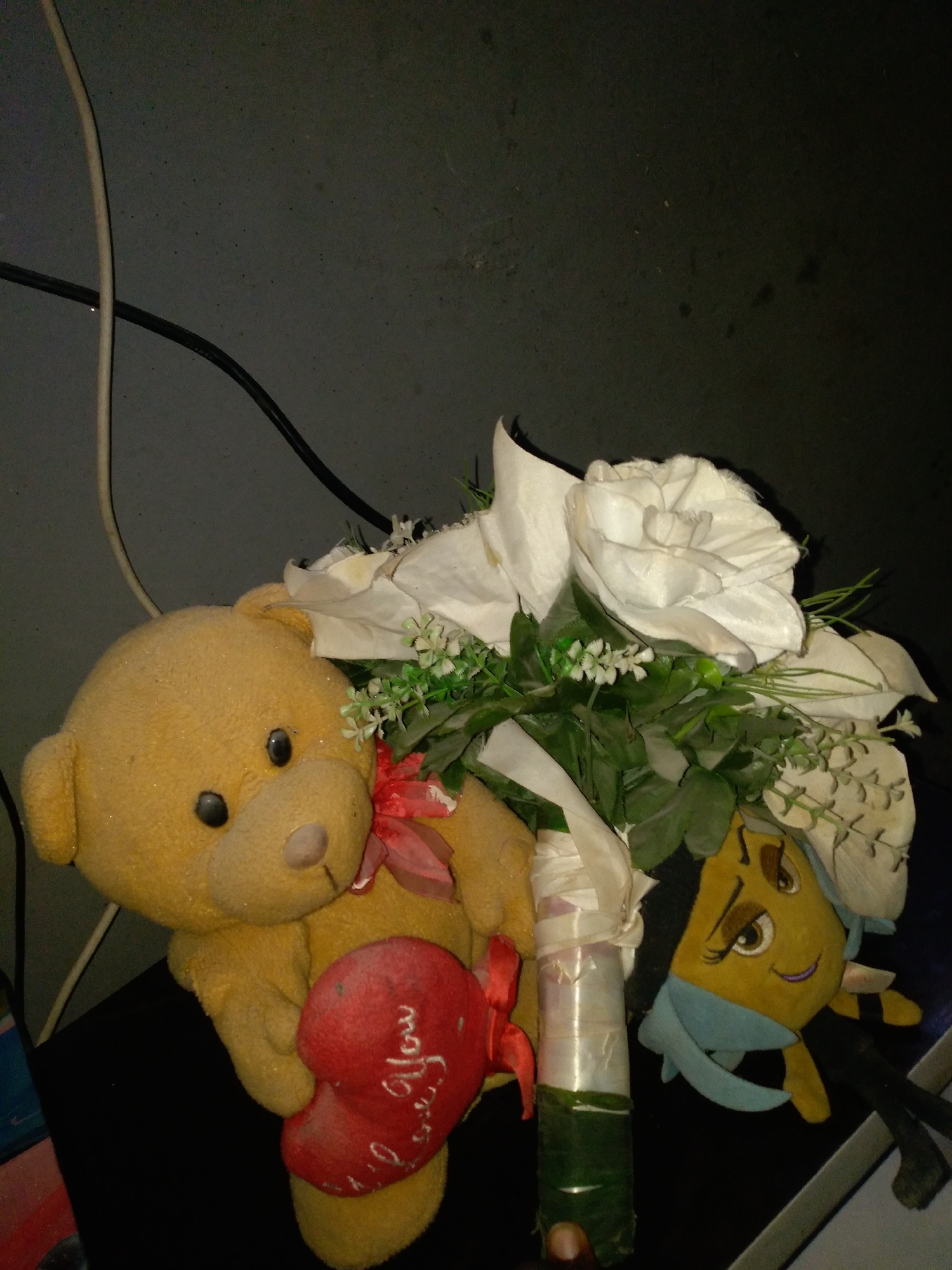 A dimly lit scene features a yellow teddy bear with black button eyes and a pink ribbon tied around its neck, holding a red heart with "I love You" written in white script. Beside the teddy bear is a bouquet of white artificial flowers with green foliage, wrapped in white and green material at the base. A small, yellow plush bee toy with large eyes and a blue scarf is partially visible to the right of the bouquet. The objects are placed on a dark surface, likely a table, with dark, textured walls in the background. Electrical cords are visible in the upper left portion of the image.