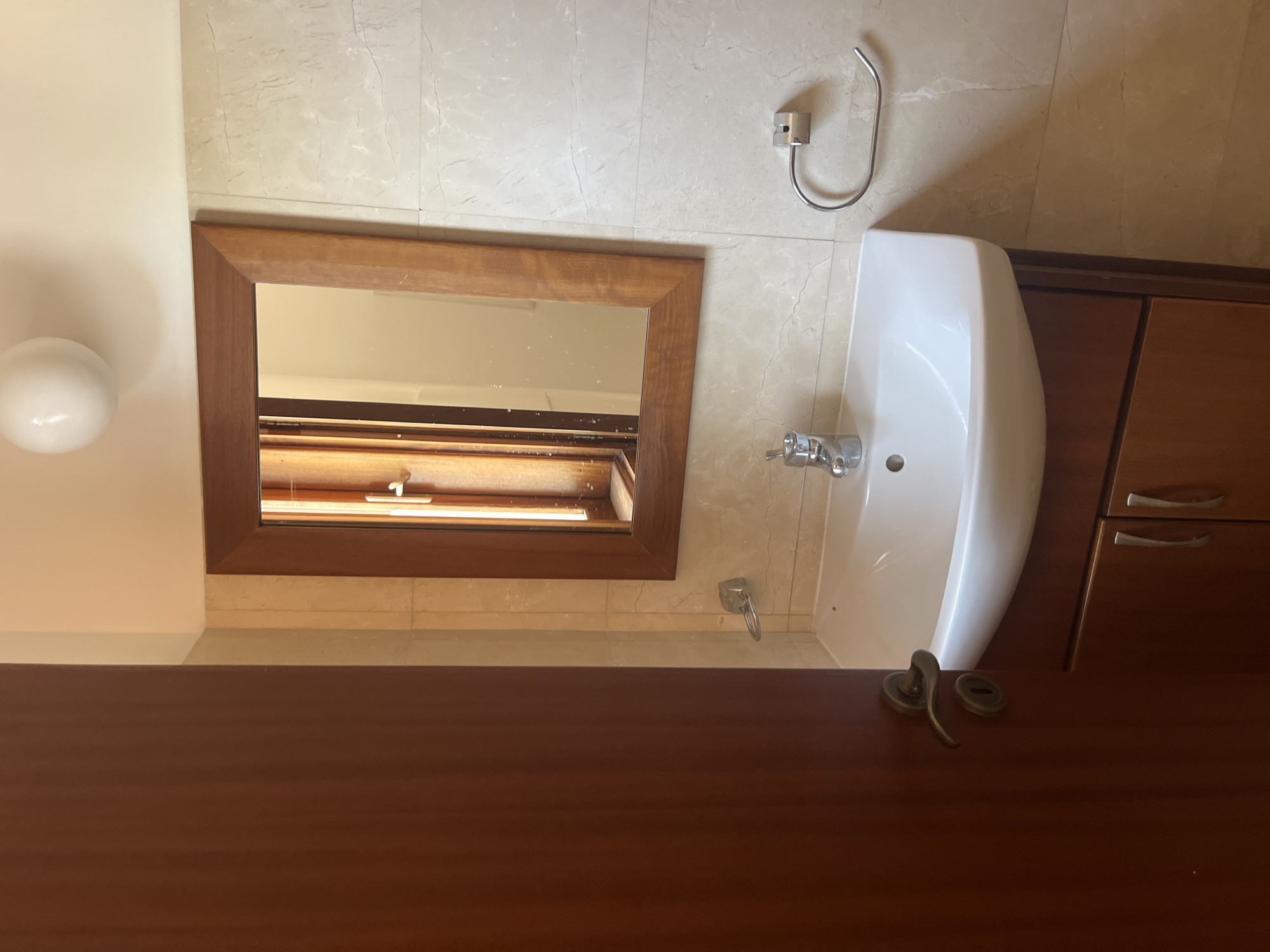 The image depicts a well-lit bathroom in a property located in Aphrodite Hills, Cyprus. The scene is dominated by a wooden-framed mirror above a white ceramic sink. The sink is set into a dark wooden vanity with two drawers. To the left of the sink, a silver soap dish is mounted on the tiled wall. A silver faucet is positioned in the center of the sink basin. On the right wall, a silver towel ring is installed. The walls are tiled with a beige, marble-like pattern, and a white spherical light fixture is visible on the ceiling. A dark brown door is partially open on the left side of the frame, revealing its handle and lock. The overall impression is of a clean and functional bathroom.