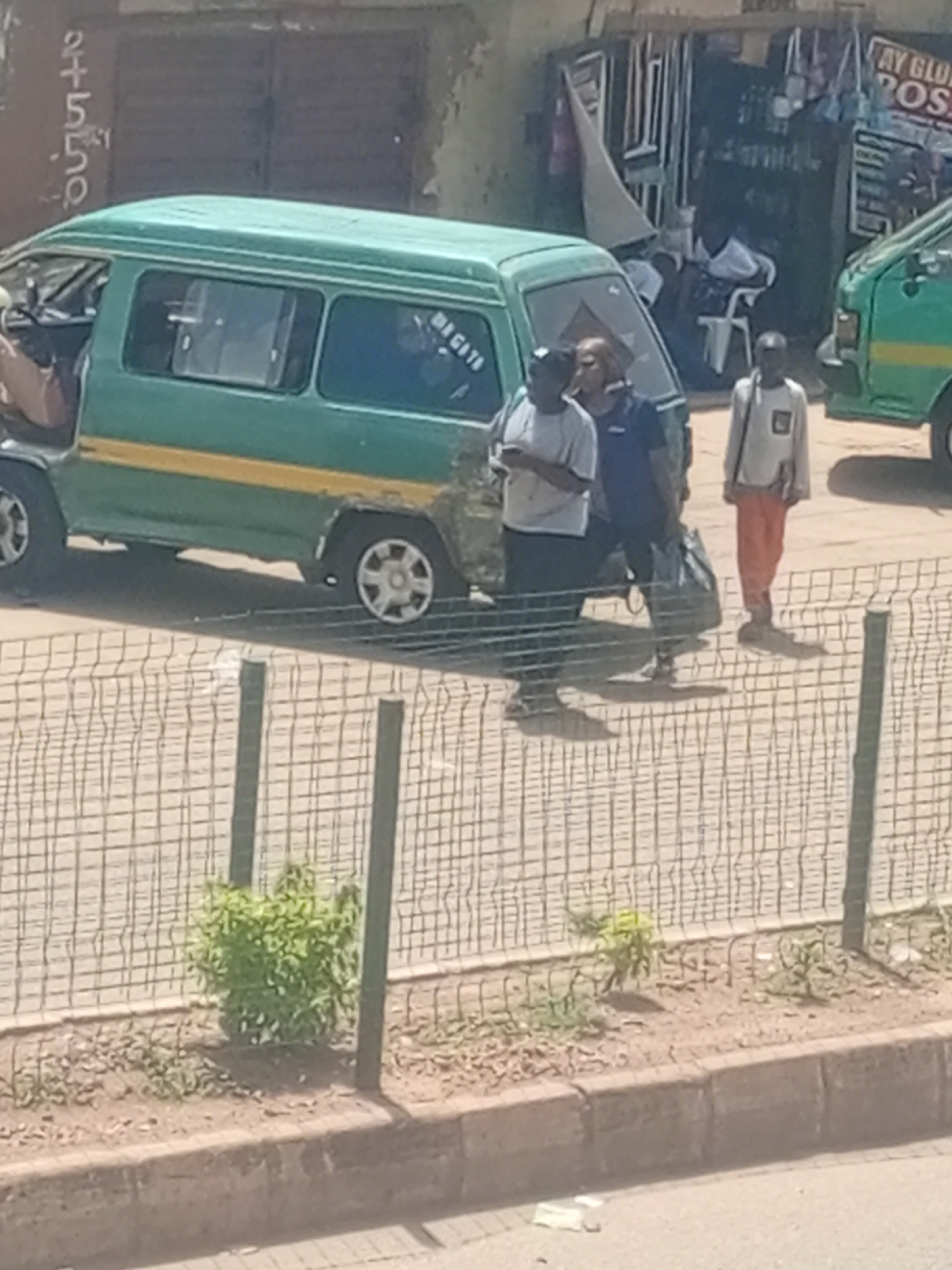 A scene unfolds in Bukuru, Nigeria, featuring a green and yellow minibus parked on a paved street. The minibus has visible signage that reads "MAGITE" on its side. To the right of the minibus, a green van is also present. In the foreground, a wire fence separates the street from a sidewalk area with sparse vegetation. Three individuals are near the minibus. One person, wearing a light-colored t-shirt and dark pants, stands to the left of the minibus, facing away from the camera. Another individual, in a dark t-shirt and dark pants, stands beside them, also facing away. A third person, dressed in a light-colored shirt and orange pants, stands further to the right, facing the camera. The background shows buildings and what appears to be a shop with various items displayed outside. The overall atmosphere suggests a typical urban street scene.