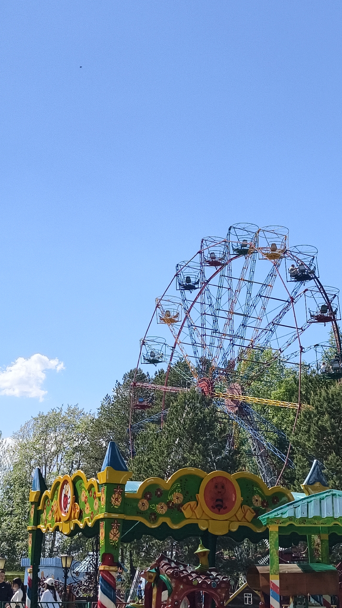 The image depicts a Ferris wheel in an amusement park in the city of Chaikovsky, Russia. The Ferris wheel has open-cage seating and is visible behind tall green trees. A colorful carousel with elaborate decorations stands in front of the Ferris wheel, partially obscuring the base. In the lower left, several people are visible near the carousel. The sky is a clear, light blue with a few small clouds.
