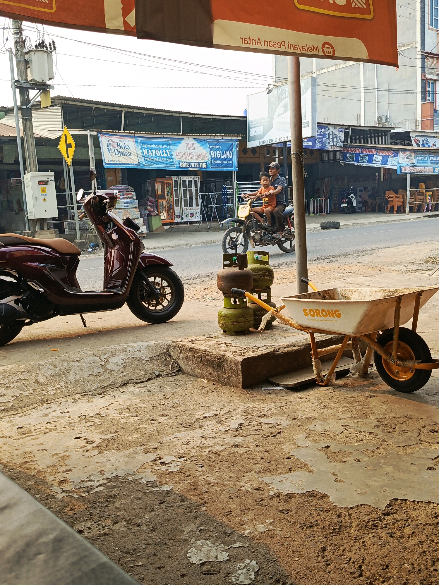 In the city of Megang Sakti II, Indonesia, a street scene unfolds with a focus on commerce and transportation. In the foreground, a maroon scooter is parked on a dusty concrete sidewalk. Next to it, a wheelbarrow with an orange frame and a white basin is situated, displaying the word "SORONG" on its side. Behind the wheelbarrow, a stack of green gas cylinders is visible, some appearing weathered.
Further down the street, a motorcycle carries two individuals: a man, presumably the driver, wearing a cap and a dark shirt, and a young boy in an orange shirt seated in front of him. They are moving away from the viewer.
The background reveals a row of shops lining the street. Banners hang above the storefronts, advertising various products and services, including mattresses, furniture, and possibly other retail goods. Electrical poles and transformers are also present, indicating the presence of infrastructure. The overall environment suggests a bustling commercial area with a mix of pedestrian and vehicular traffic, common in Indonesian street scenes. The sunlight casts shadows, indicating it is likely daytime.