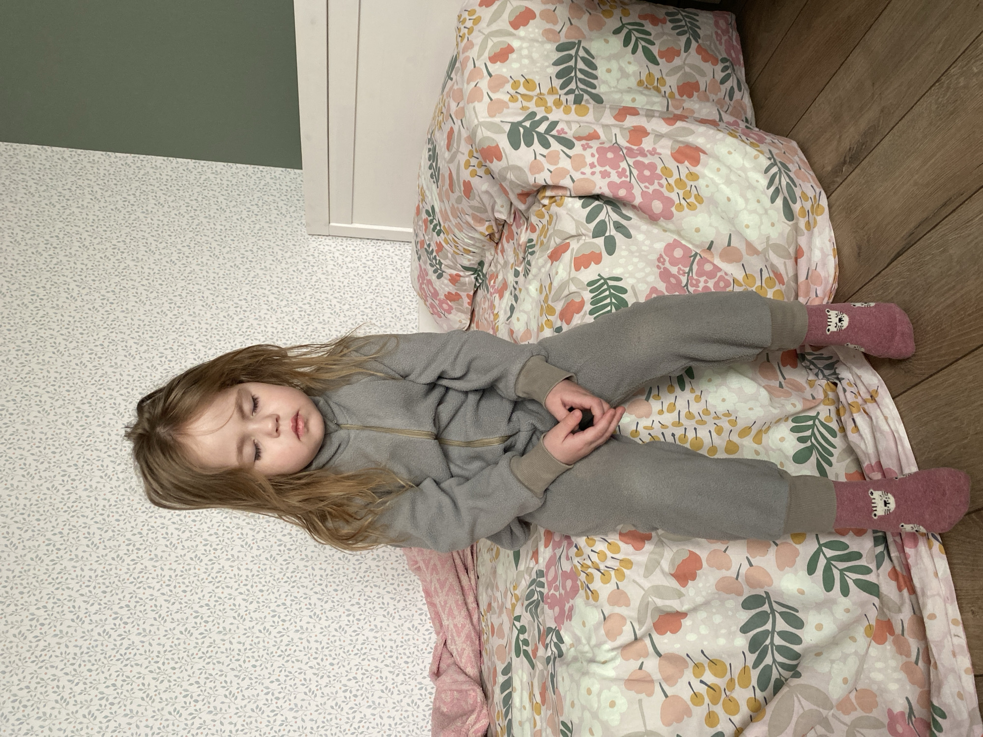 A young child with long blonde hair, wearing a gray zip-up jumpsuit and patterned pink socks, is lying on a bed covered with a floral patterned comforter and a light pink blanket. The child is asleep with closed eyes and clasped hands. The bed is positioned against a wall with a white door frame and patterned wallpaper with small, repetitive leaf-like designs. Above the wallpaper, the wall is painted a solid green color. The floor is made of wooden planks. The location is unspecified; there is no visible evidence to determine if the photo was taken in Izhevsk, Russia.